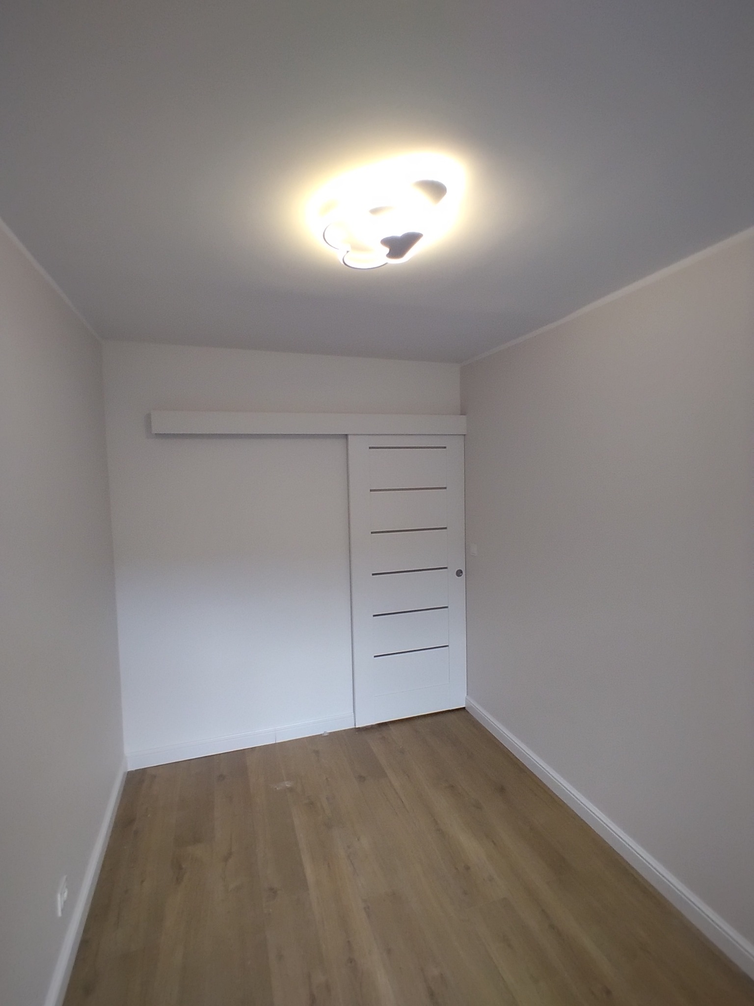 Wnętrze pokoju po remoncie: ułożone panele podłogowe w odcieniach brązu, białe ściany, przesuwne drzwi z poziomymi wgłębieniami i nowoczesna lampa sufitowa.