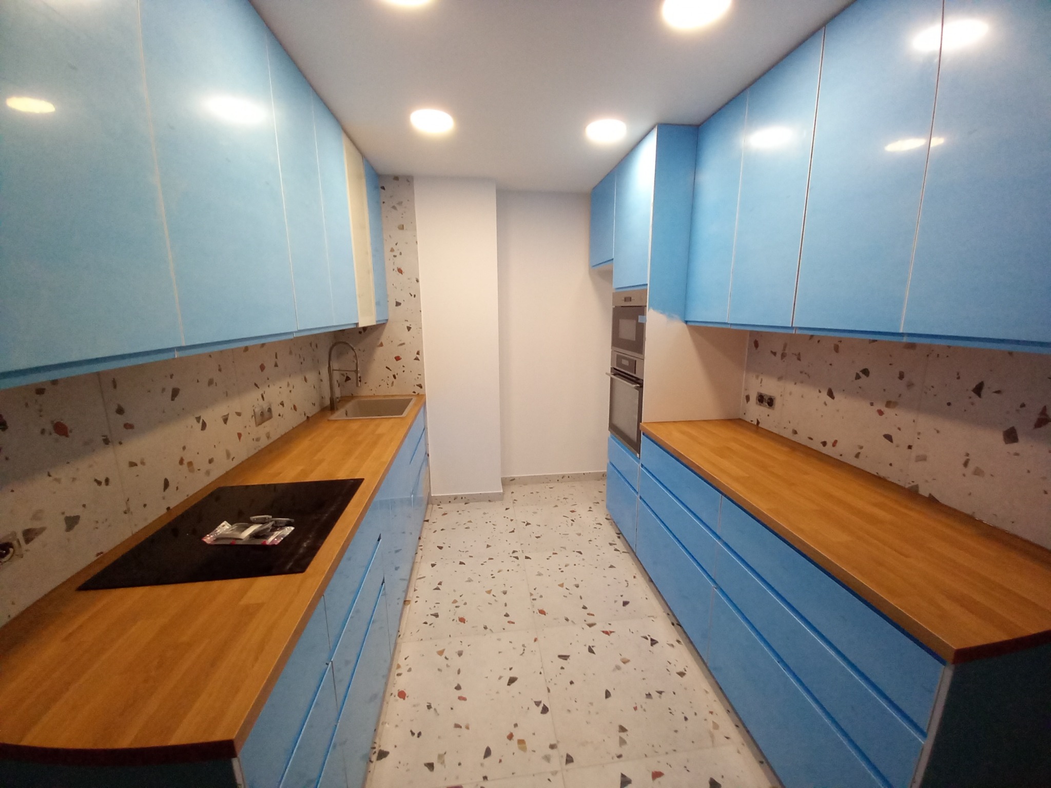 Wąska kuchnia z błękitnymi szafkami i drewnianymi blatami, terazzo na podłodze i ścianie nad blatem, płyta indukcyjna na blacie po lewej stronie.