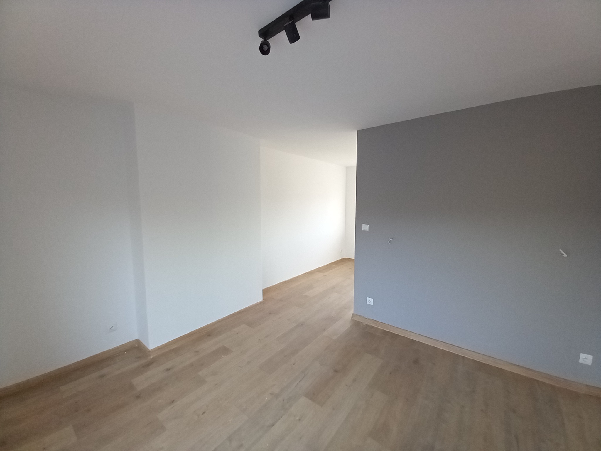 Pusty pokój po remoncie z panelami podłogowymi w odcieniach jasnego drewna i świeżo pomalowanymi ścianami, jedna ściana w kolorze szarym, sufit biały z reflektorami.