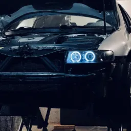 Srebrny samochód z otwartą maską, podniesiony na podnośniku; widoczne okrągłe, niebieskie światła LED w reflektorach.