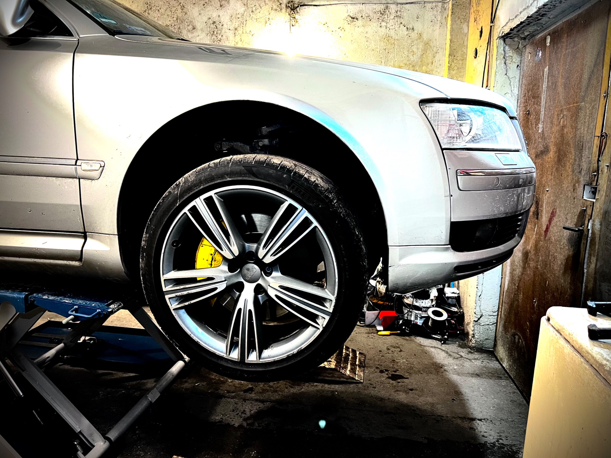 Srebrny samochód na podnośniku w warsztacie, widoczne koło z żółtym zaciskiem hamulcowym, narzędzia na podłodze.