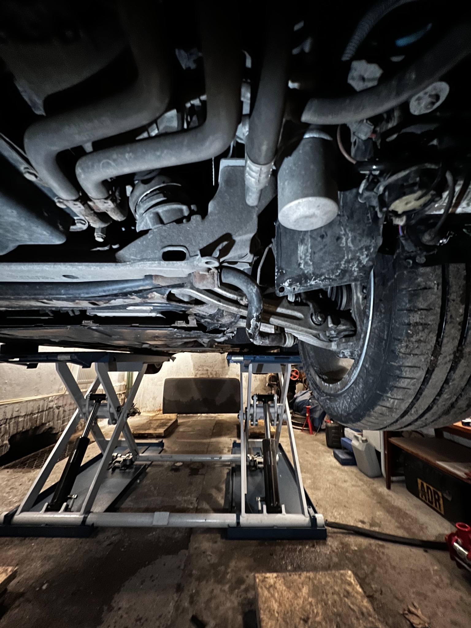 Widok podwozia samochodu na podnośniku nożycowym, widoczne elementy zawieszenia, układu wydechowego i koło z oponą. Niska perspektywa, garażowe otoczenie.