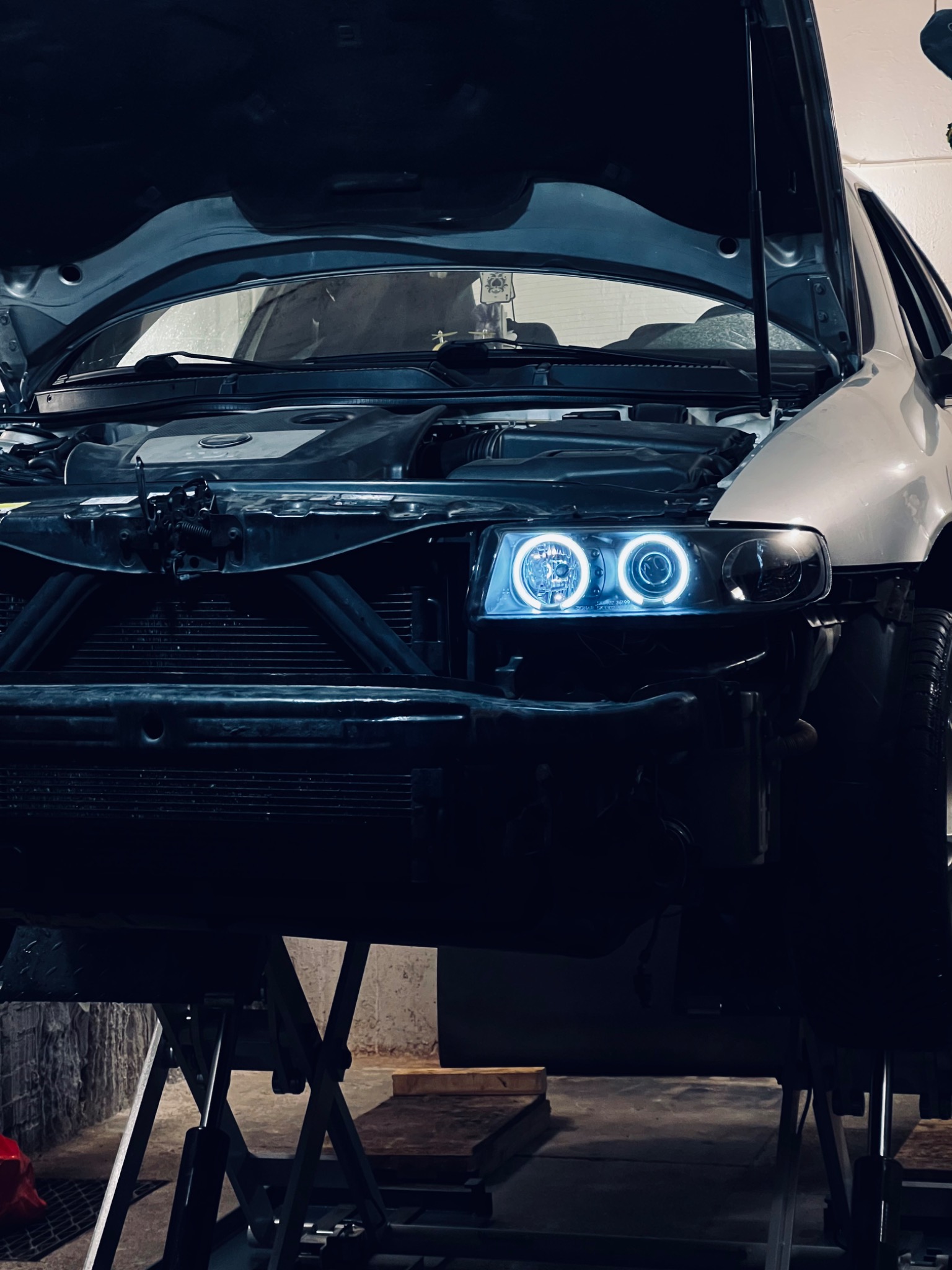 Srebrny samochód z otwartą maską, podniesiony na podnośniku; widoczne okrągłe, niebieskie światła LED w reflektorach.