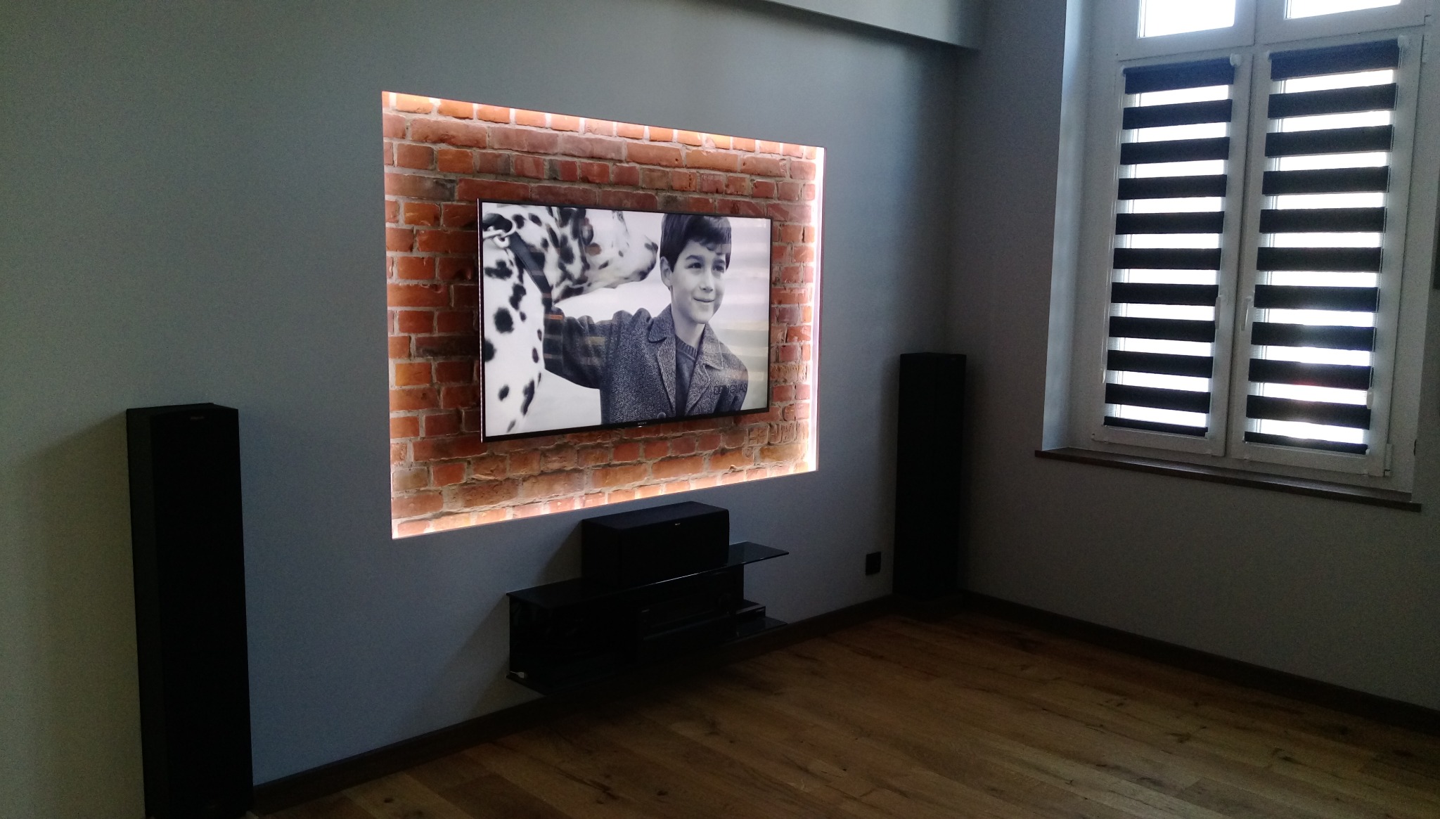 Salon z telewizorem w obudowie z cegły z podświetleniem LED, czarne głośniki podłogowe po bokach i drewniana podłoga.