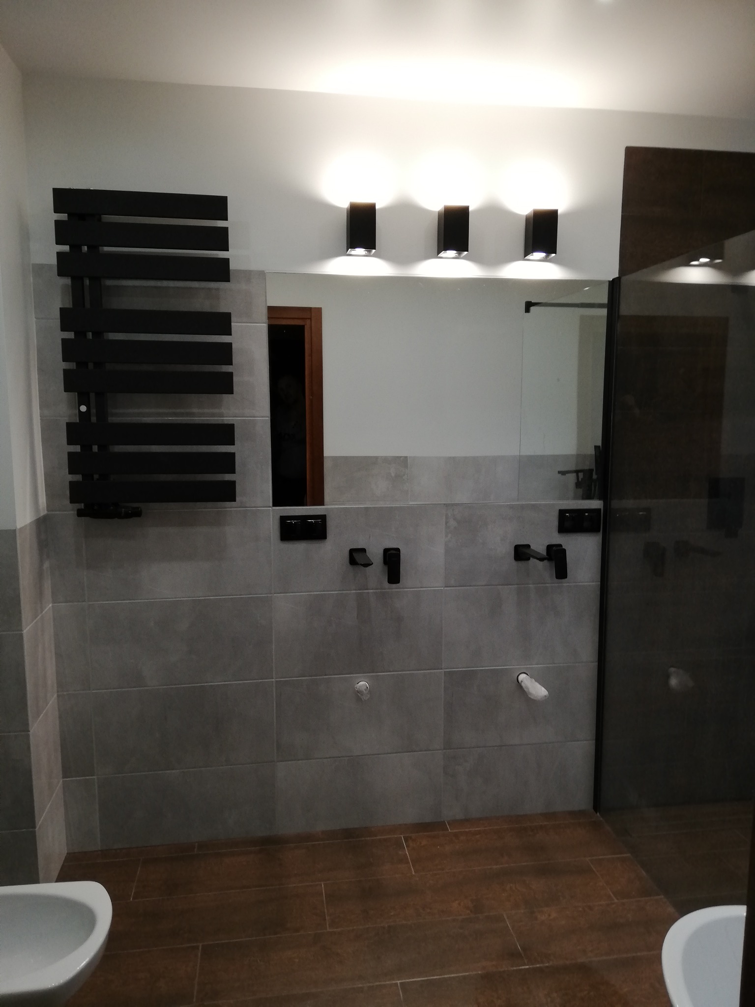 Nowoczesna łazienka z szarymi płytkami, drewnopodobną podłogą, czarną armaturą i grzejnikiem drabinkowym, oświetlona trzema kinkietami nad lustrem.