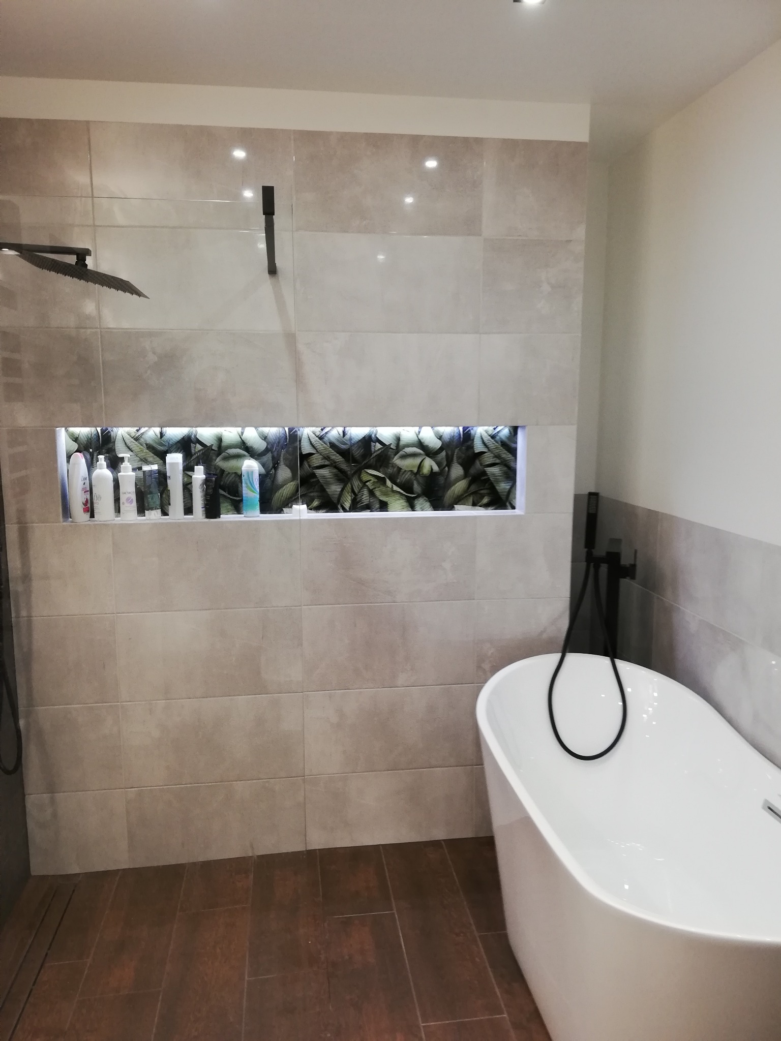 Nowoczesna łazienka z wolnostojącą białą wanną, ciemną armaturą i wnęką w ścianie wyłożonej beżowymi płytkami, podświetloną i wypełnioną kosmetykami na tle dekoracyjnej tapety z motywem liści.