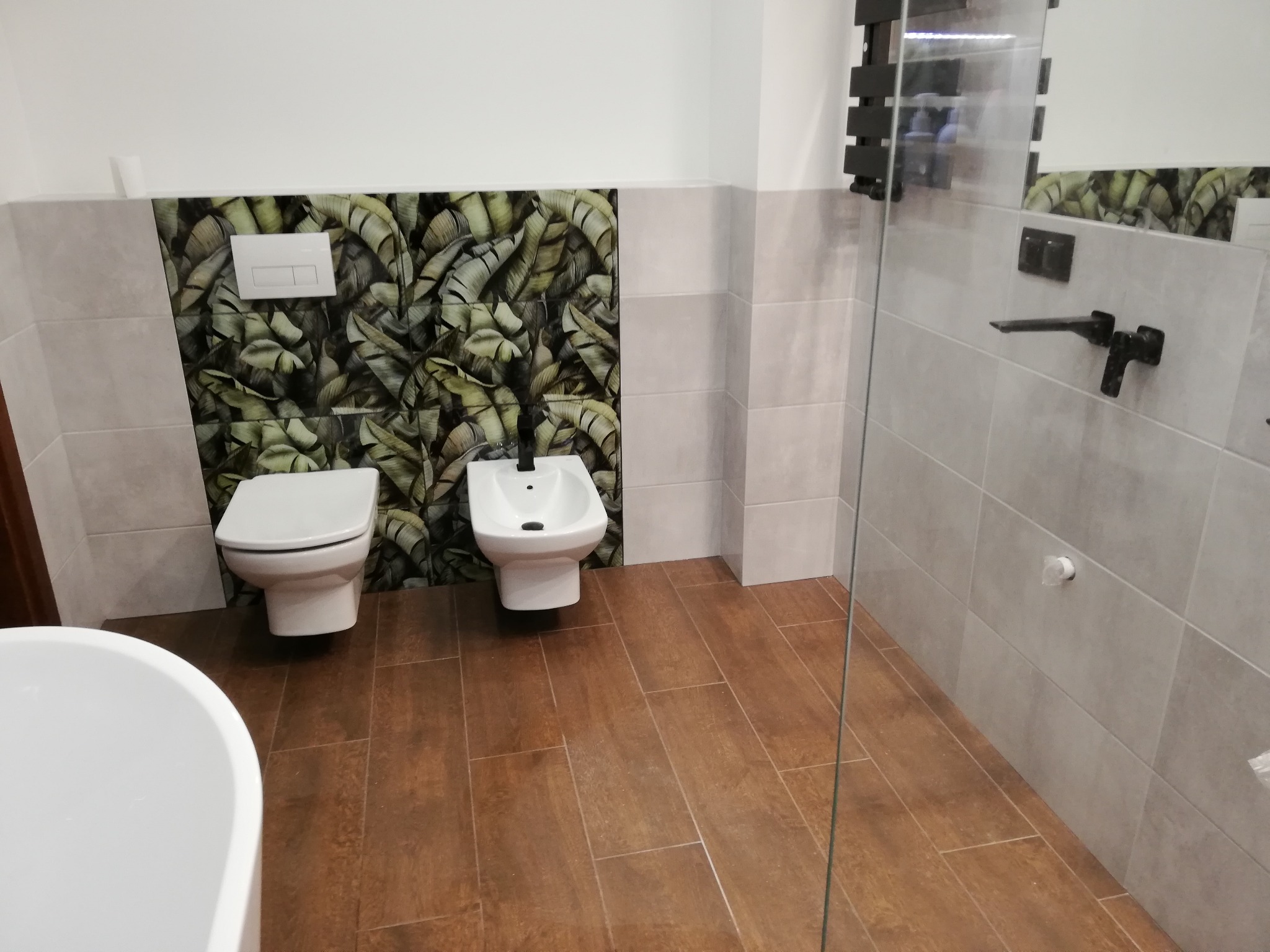 Nowoczesna łazienka z brązowymi płytkami podłogowymi imitującymi drewno, szarymi płytkami ściennymi, toaletą i bidetem na tle ściany z motywem liści bananowca, widoczna częściowo wanna.