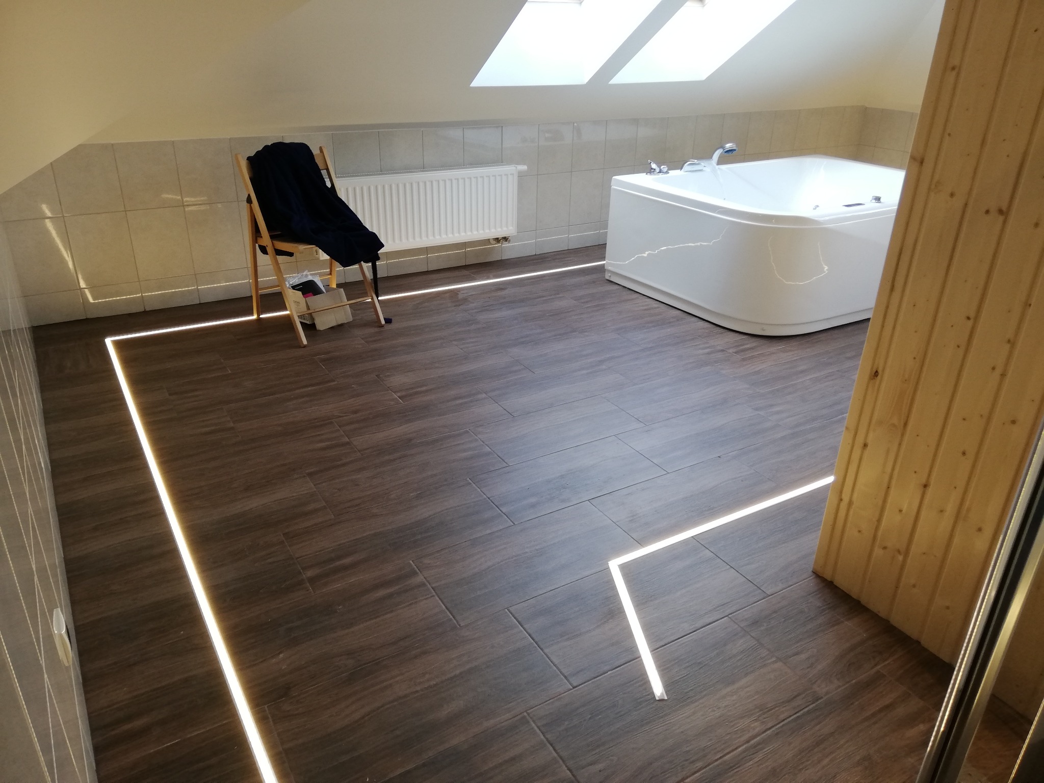 Podświetlona podłoga z ciemnych paneli imitujących drewno w łazience na poddaszu, biała wanna narożna, krzesło z ubraniem i grzejnik na tle jasnych płytek, oświetlenie LED wzdłuż krawędzi ścian.