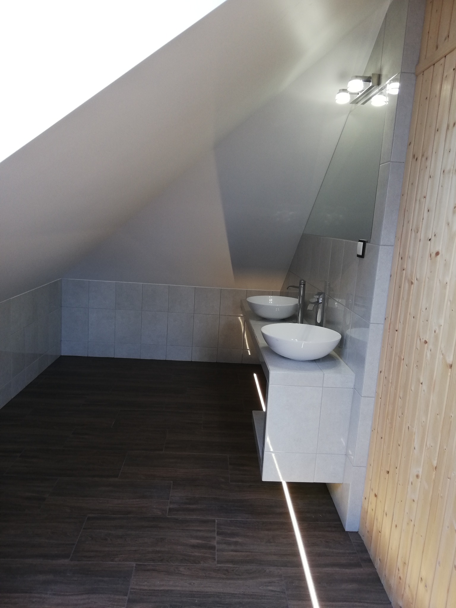 Nowoczesna łazienka na poddaszu z dwoma umywalkami nablatowymi w kształcie misy, szarą glazurą ścienną, podświetlaną szafką pod umywalkami i ciemną podłogą imitującą drewno.