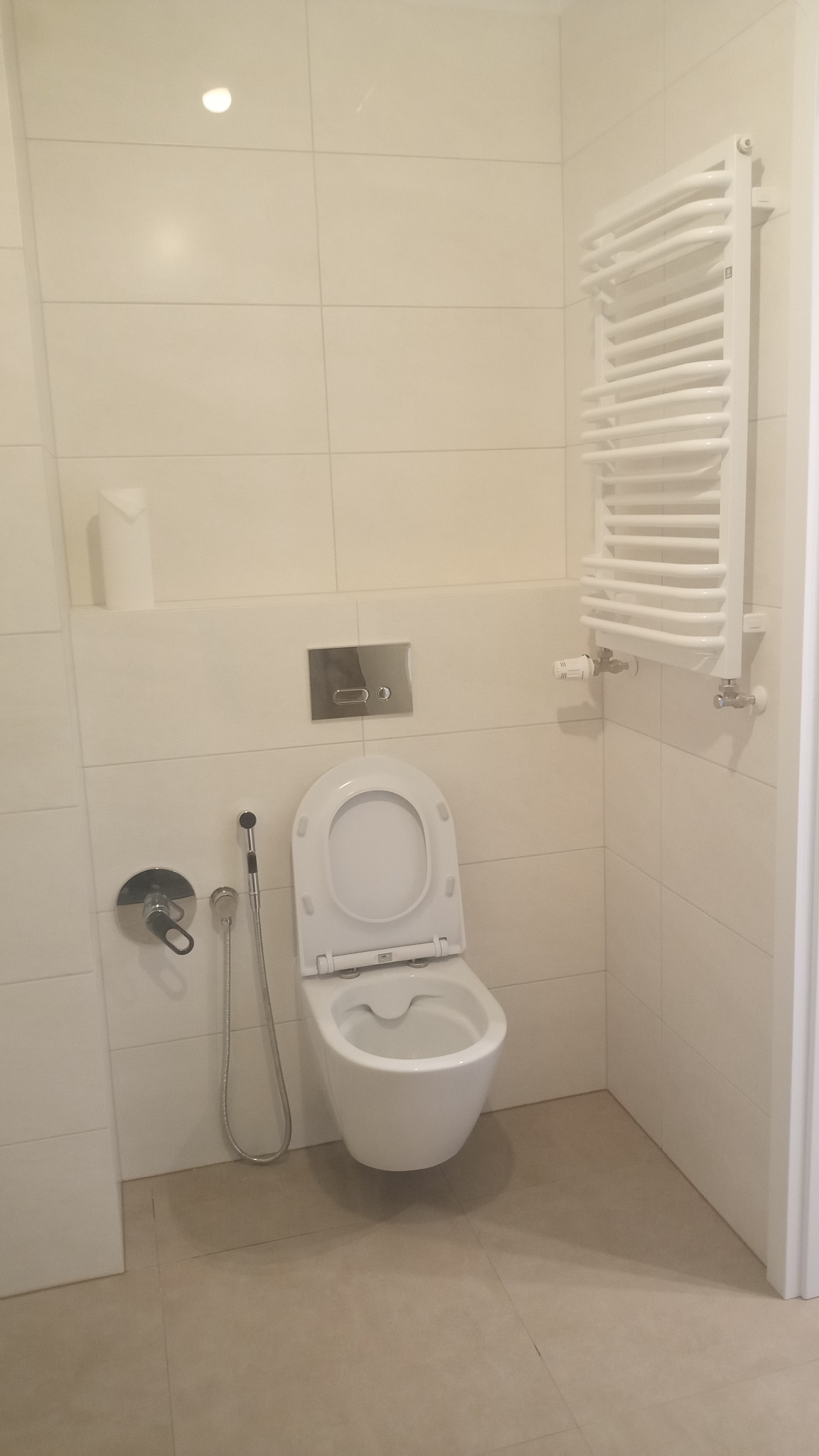 Nowoczesna, minimalistyczna łazienka z wiszącą toaletą z otwartą klapą, bidetką, chromowaną armaturą i białym grzejnikiem drabinkowym na tle jasnych, kwadratowych płytek ceramicznych.