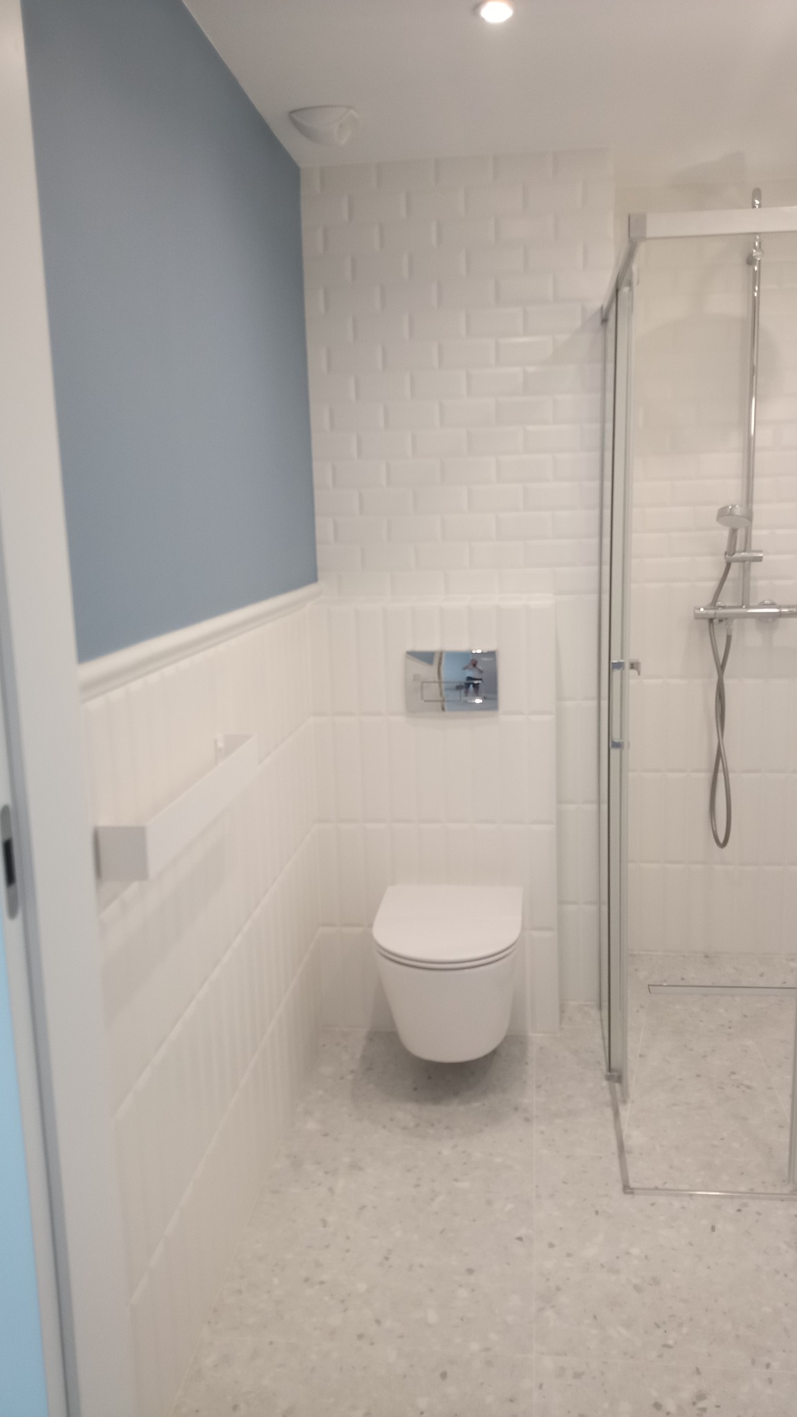 Nowoczesna łazienka z wiszącą toaletą, kabiną prysznicową ze szklanymi drzwiami, białą mozaikową glazurą ścienną i niebieską ścianą akcentującą.