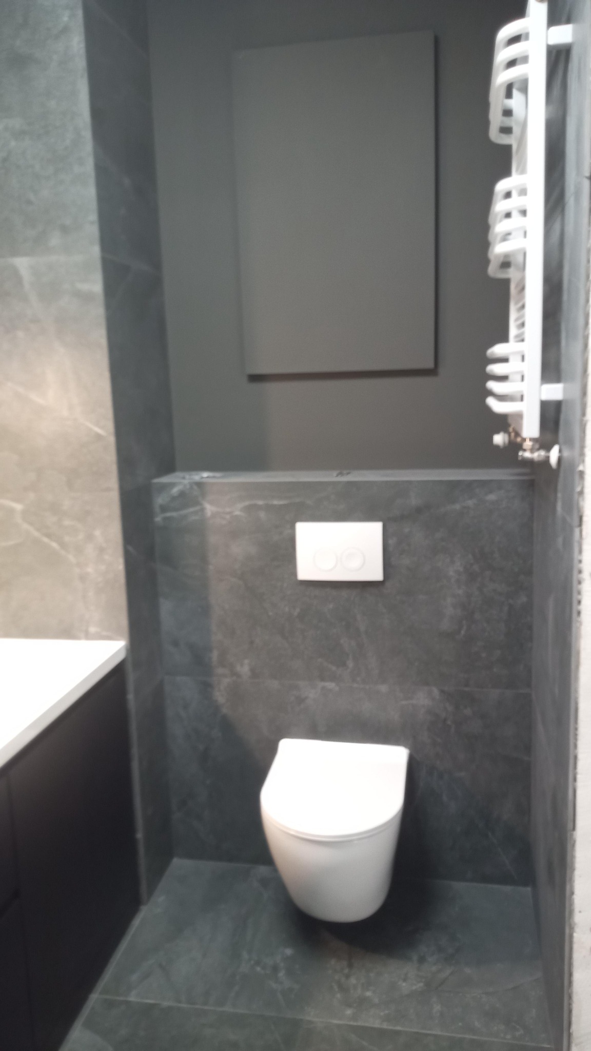Podwieszana toaleta w łazience wykończonej szarym marmurem, z widocznym grzejnikiem drabinkowym i panelem ściennym w odcieniu grafitu.