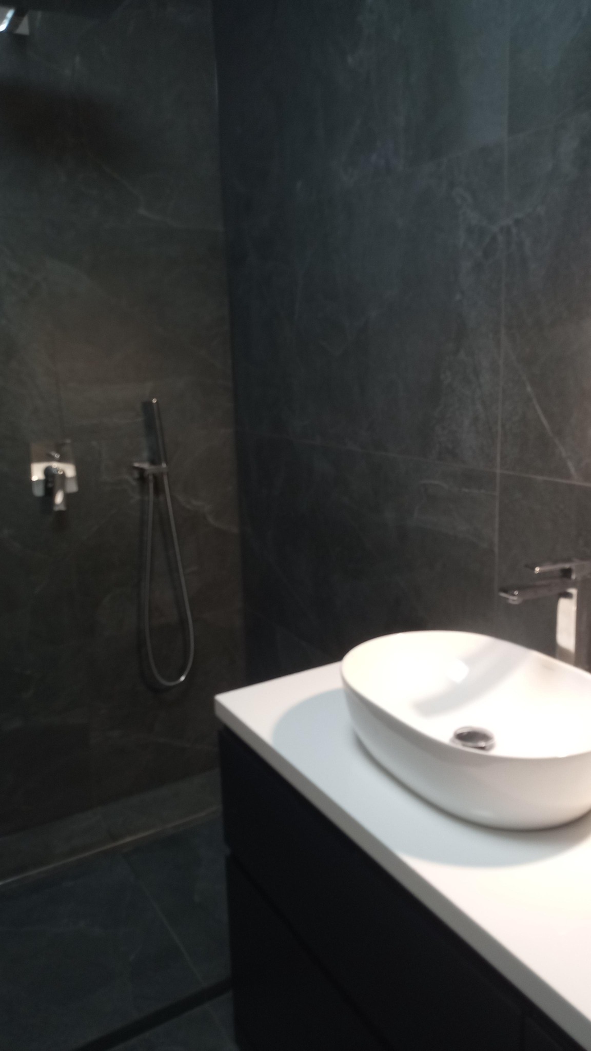 Nowoczesna łazienka z ciemnymi płytkami imitującymi marmur, białą owalną umywalką nablatową na czarnej szafce, chromowaną armaturą i deszczownicą.