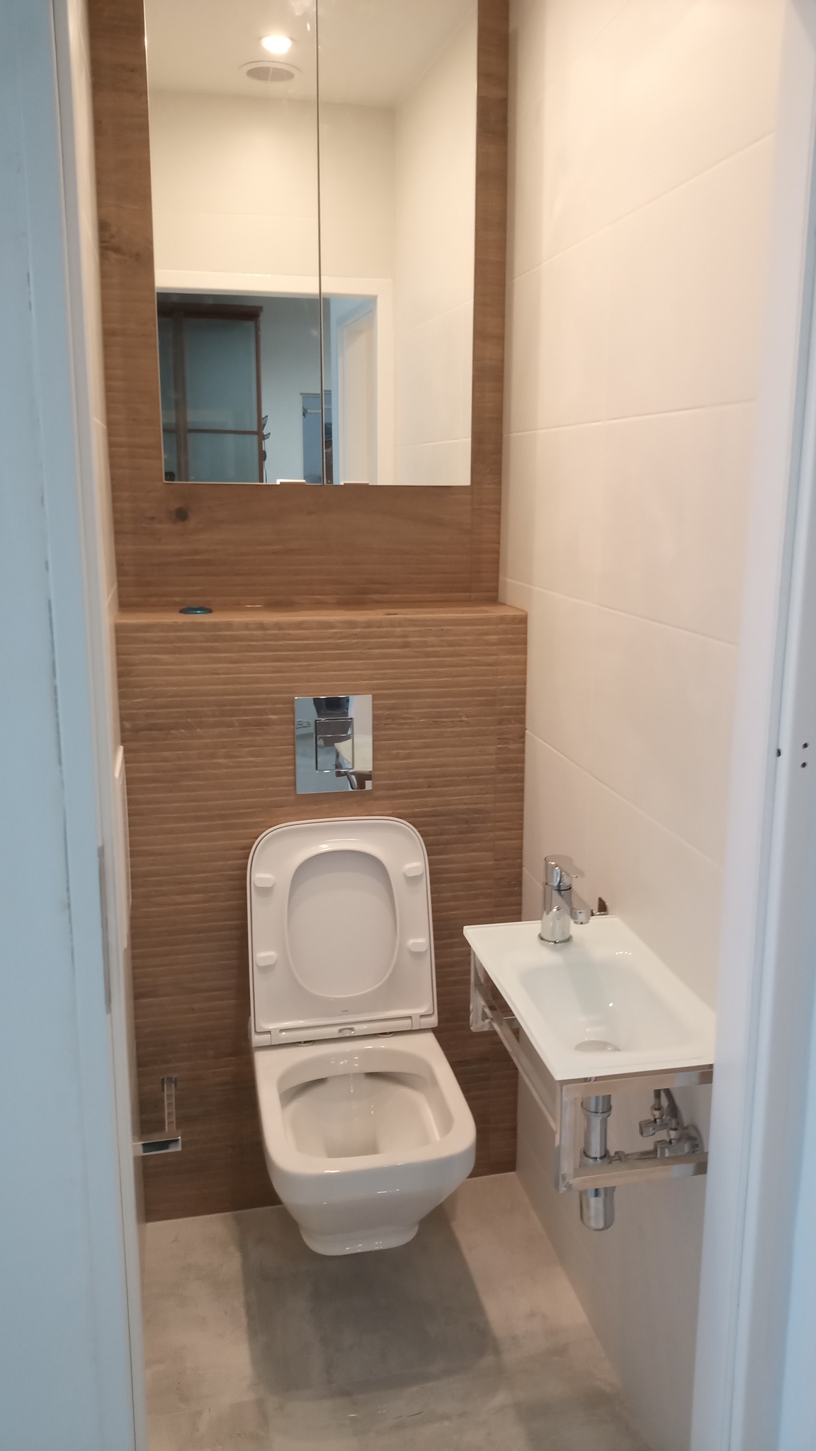 Nowoczesna łazienka z podwieszaną toaletą i umywalką, drewnopodobne panele ścienne za toaletą, białe płytki na ścianie za umywalką, lustro z drewnianą ramą.