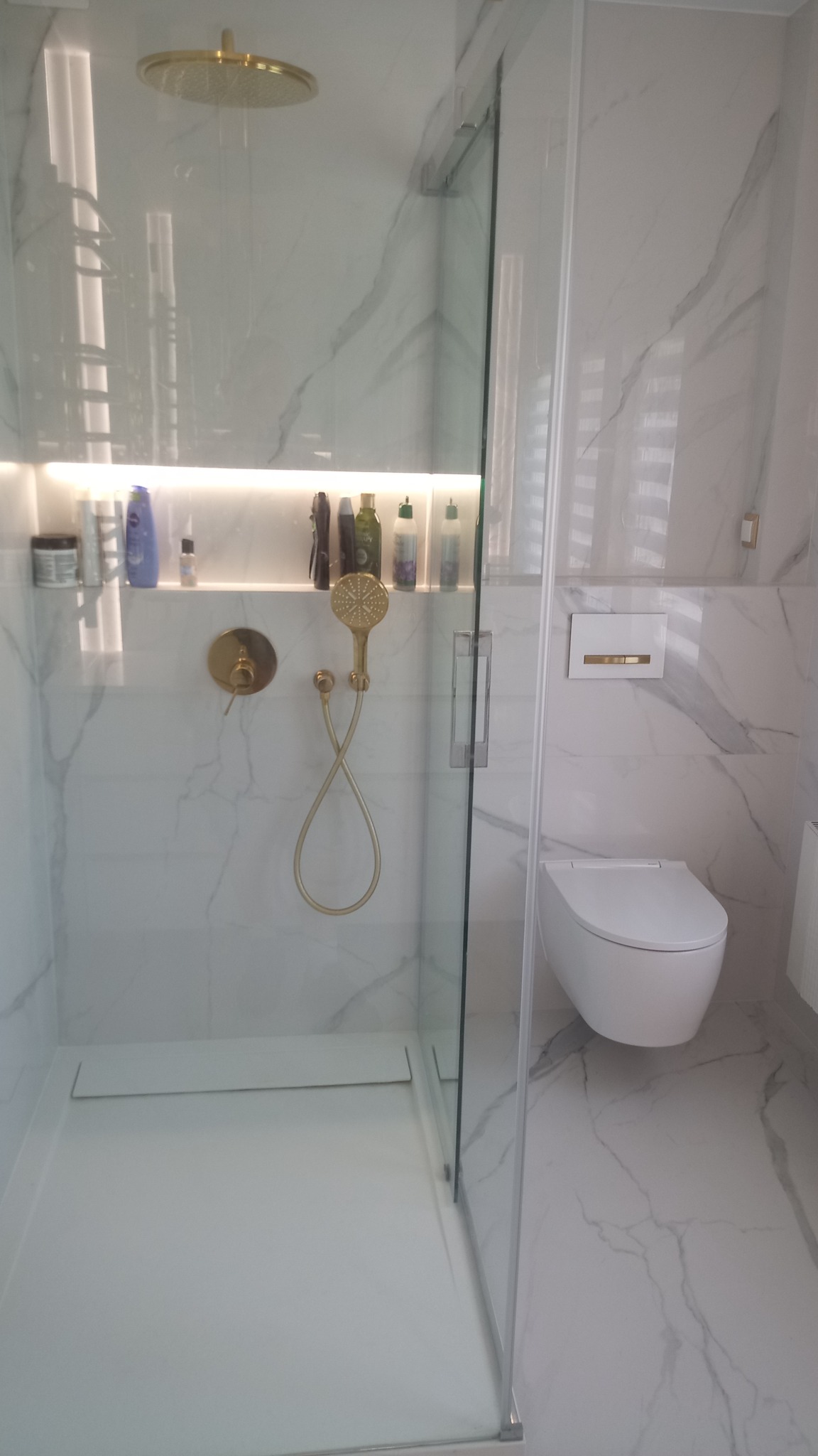 Nowoczesna kabina prysznicowa z deszczownicą i złotą armaturą, białe marmurowe płytki na ścianach i podłodze, wisząca toaleta.