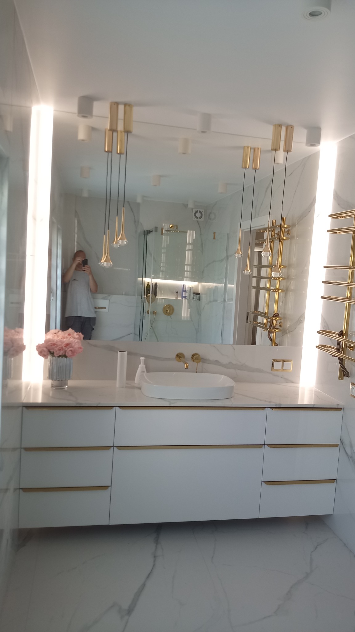 Nowoczesna łazienka z białymi szafkami pod umywalką, marmurowymi ścianami i złotymi akcentami, w tym wiszącymi lampami i grzejnikiem drabinkowym; odbicie fotografa w lustrze.