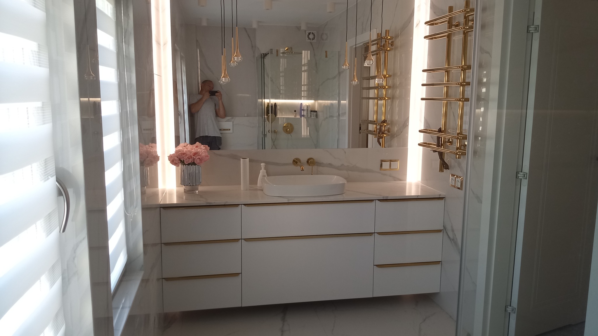 Nowoczesna łazienka z białymi szafkami z złotymi uchwytami, białą umywalką nablatową, marmurowym blatem i ścianą oraz złotym grzejnikiem drabinkowym, widoczne odbicie fotografa w lustrze.