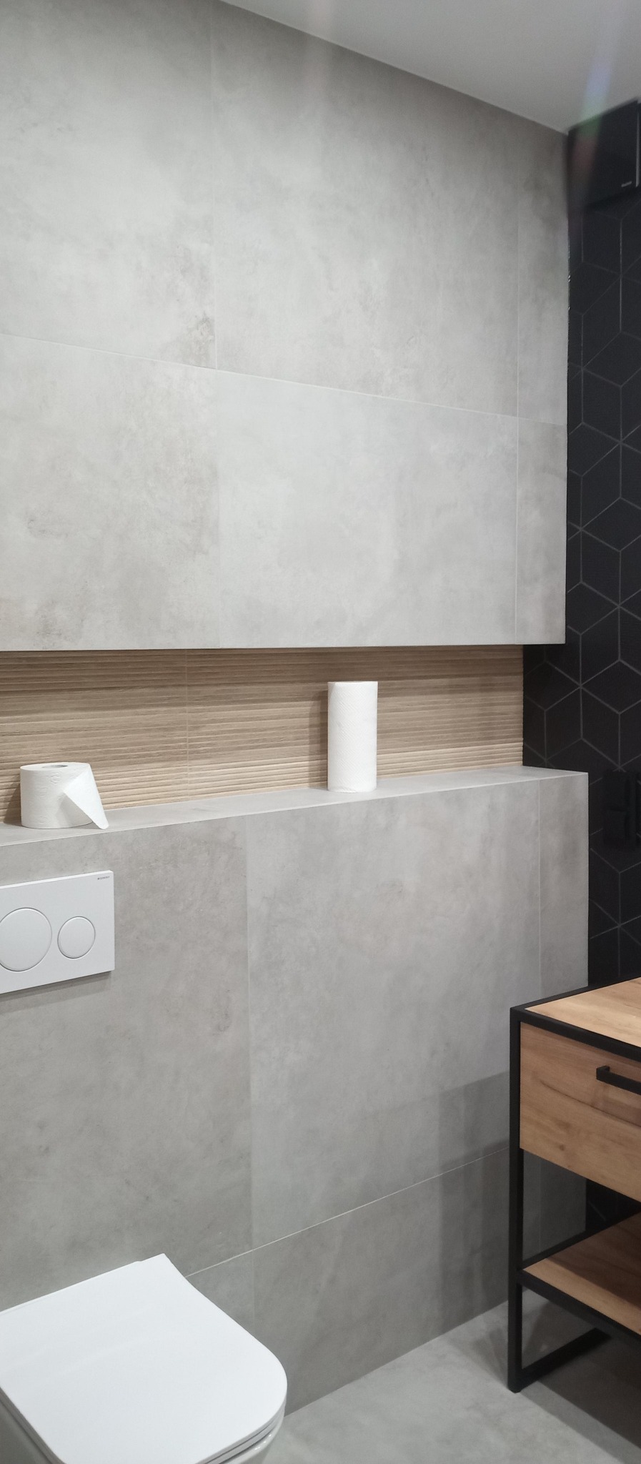 Wykończona łazienka z szarymi płytkami imitującymi beton, drewnianą półką z rolkami papieru toaletowego, wiszącą toaletą i szafką z drewnianym frontem i czarnymi metalowymi nogami.