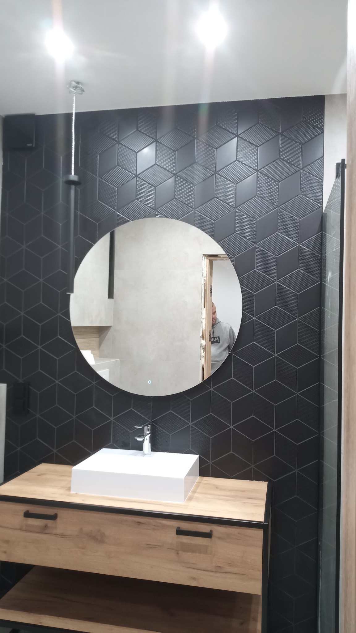 Nowoczesna łazienka w Gdańsku z czarnymi płytkami o geometrycznym wzorze 3D, okrągłe lustro z podświetleniem LED, biała umywalka nablatowa na drewnianej szafce z czarnymi uchwytami.