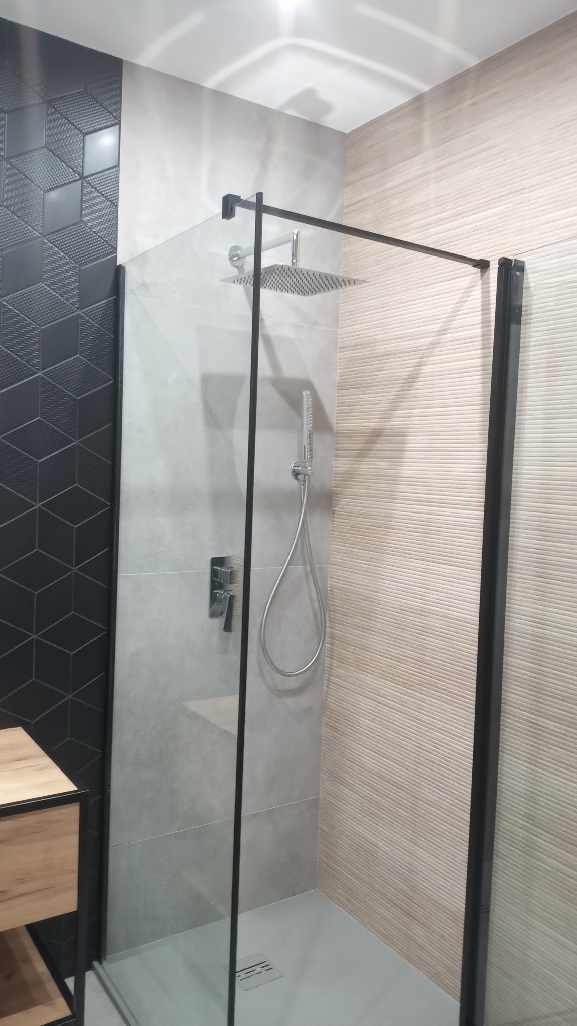Nowoczesna kabina prysznicowa z czarnymi ramami, deszczownicą i baterią podtynkową, wykończona płytkami w odcieniach szarości i beżu, z elementami dekoracyjnymi w geometryczny wzór.