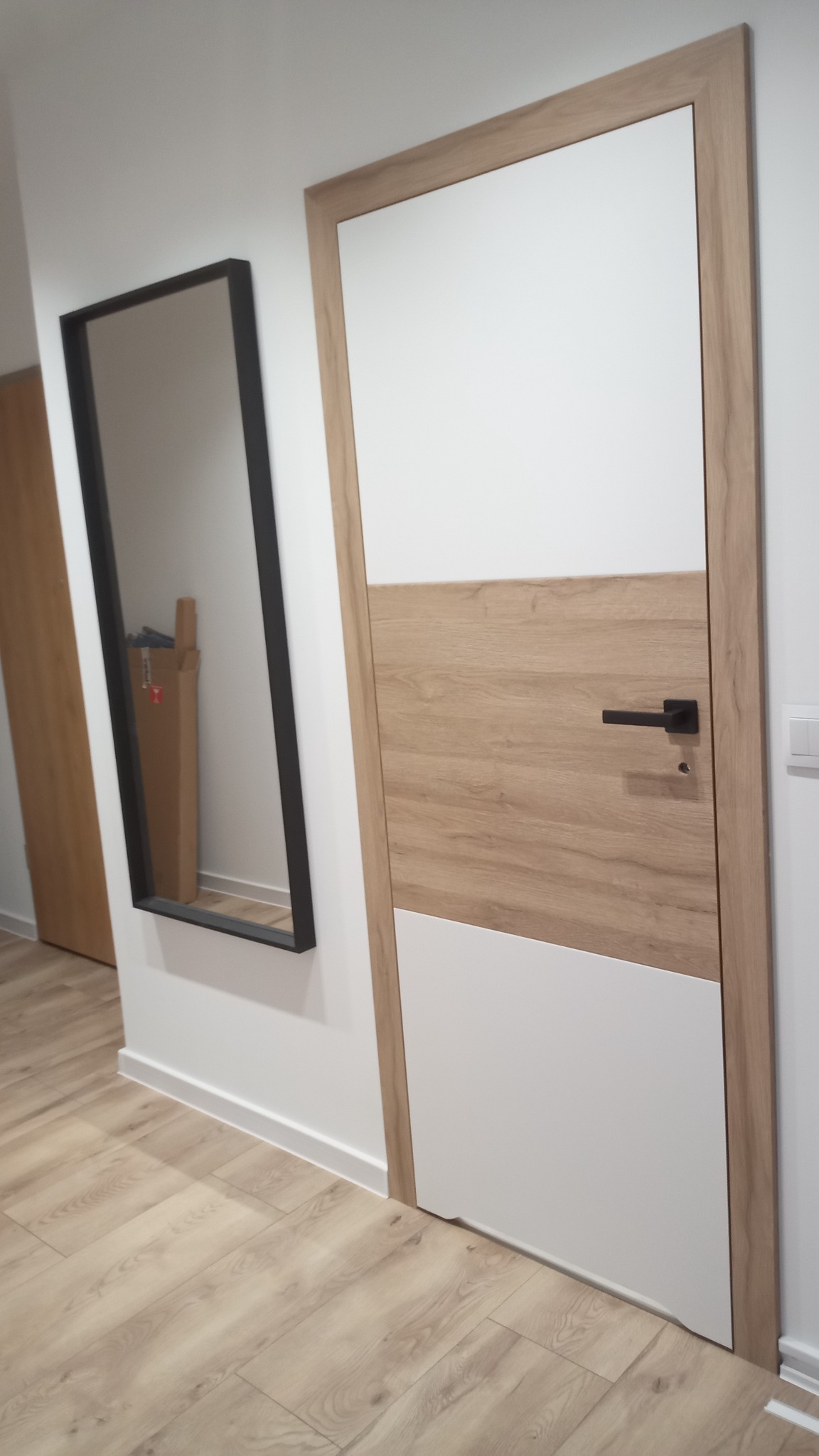Nowoczesne drzwi wewnętrzne z drewnianą ramą i panelami w kolorze drewna i bieli, czarna klamka, wiszące lustro w czarnej ramie odbijające kartonowe pudełko, panele podłogowe.