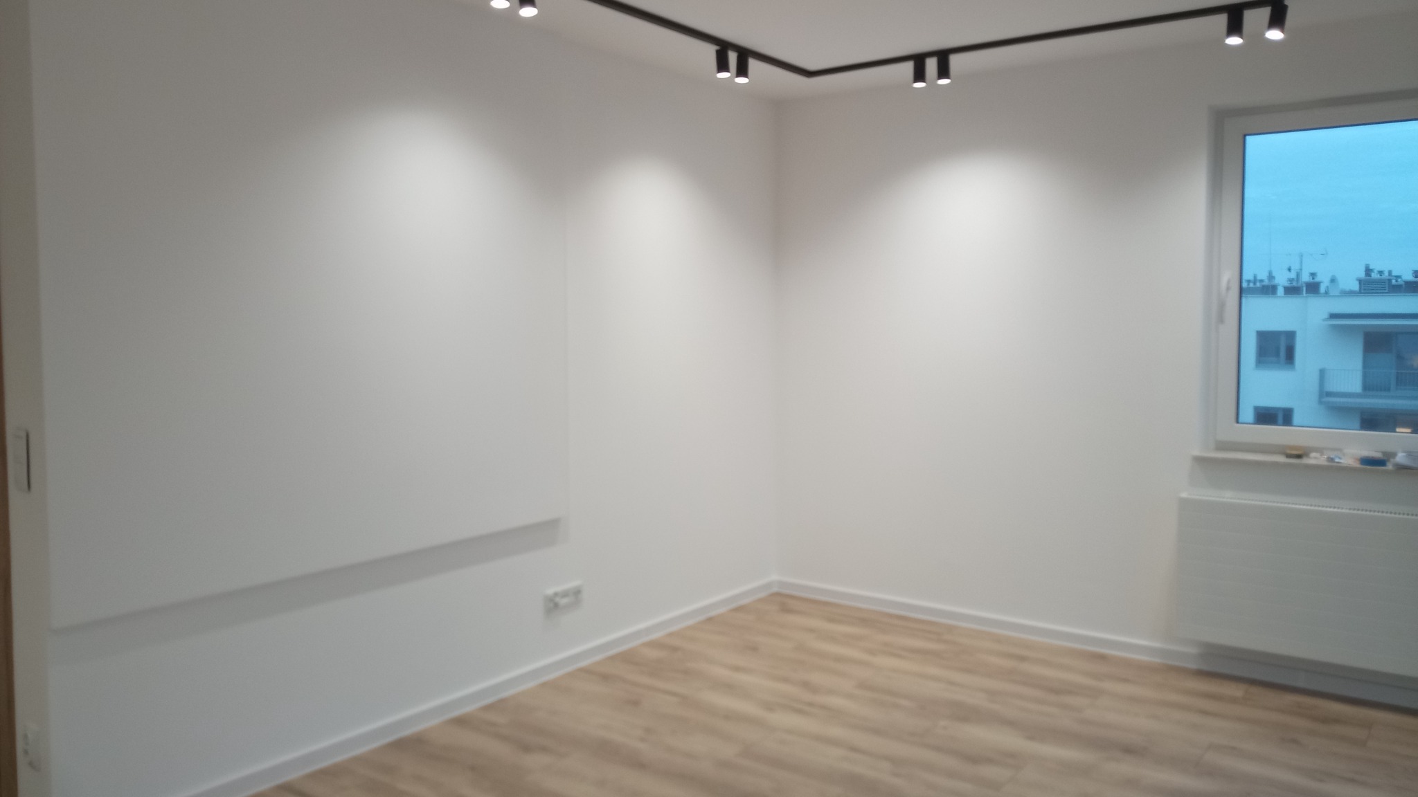 Wnętrze pokoju po remoncie: białe ściany, panele podłogowe w odcieniach drewna, nowoczesne oświetlenie punktowe na szynoprzewodzie, grzejnik pod oknem z widokiem na budynek mieszkalny.