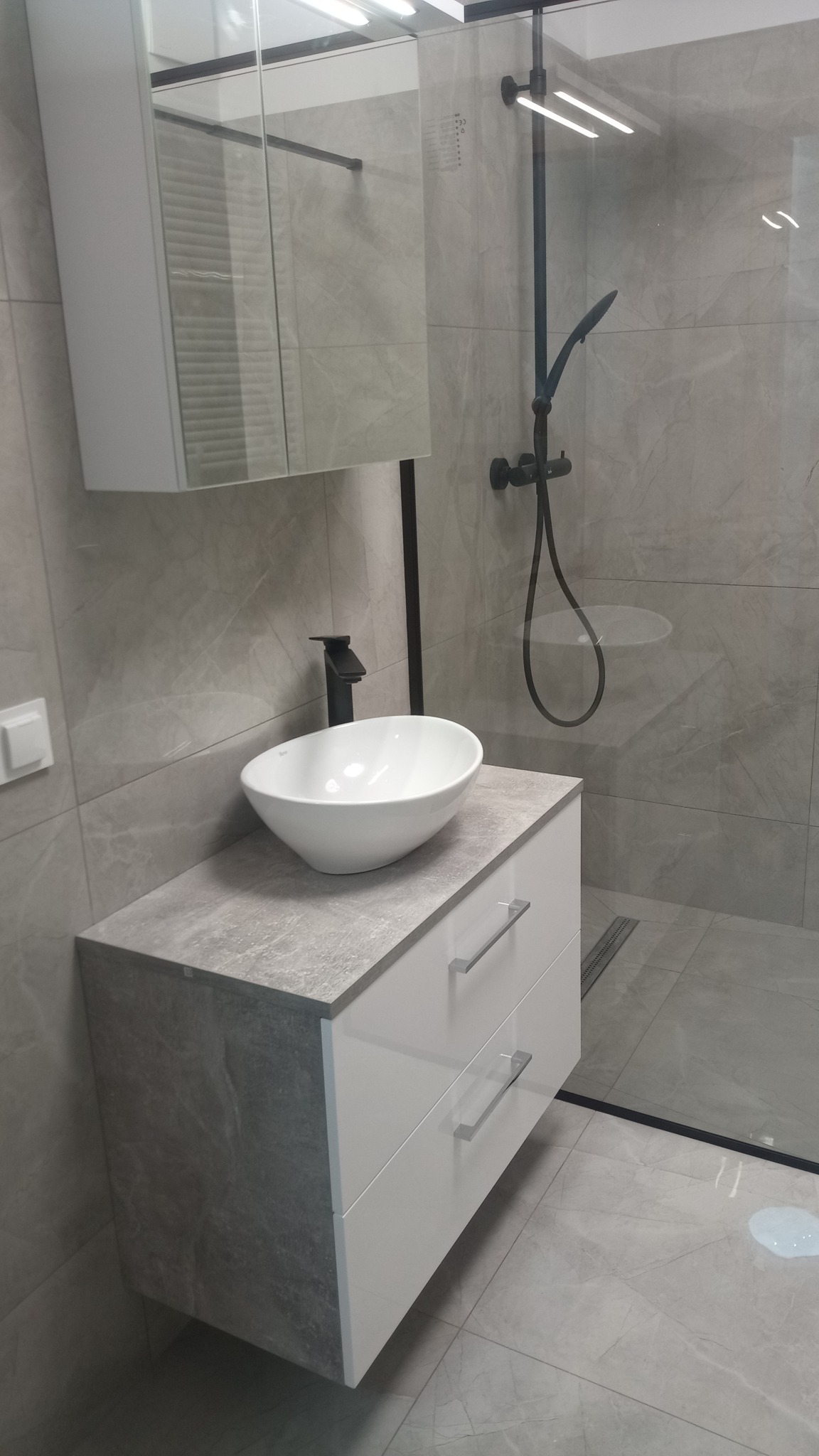 Nowoczesna łazienka z umywalką nablatową w kształcie misy, czarną armaturą i kabiną prysznicową walk-in z odpływem liniowym, wykończona szarymi płytkami imitującymi marmur.