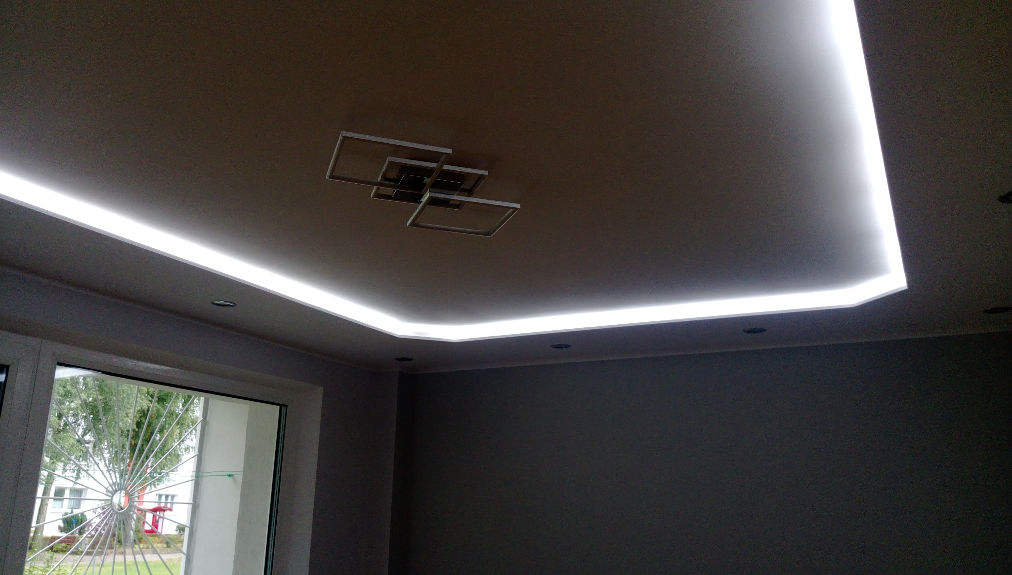 Sufit podwieszany z oświetleniem LED wzdłuż krawędzi, nowoczesna lampa sufitowa w geometryczne kwadraty, szara ściana, okno z widokiem na zieleń.