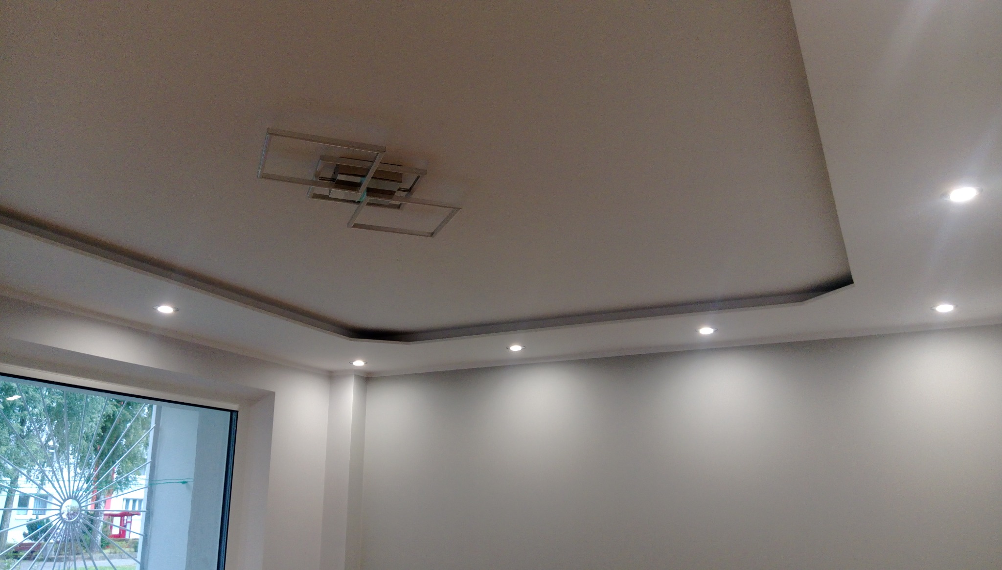 Wnętrze z pomalowanymi na biało ścianami, podwieszanym sufitem z oświetleniem punktowym i nowoczesną lampą sufitową z geometrycznych elementów, widok z dołu.