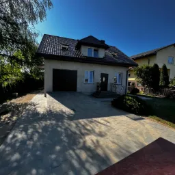 Dwukondygnacyjny dom jednorodzinny z garażem i podjazdem wyłożonym szarą kostką brukową, oświetlony mocnym słońcem, z widocznymi cieniami drzew.