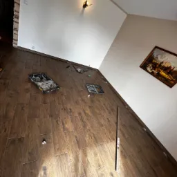 Ukośnie ułożone, brązowe płytki podłogowe imitujące drewno, z rozłożonymi narzędziami i otwartą skrzynką narzędziową, widoczne fragmenty białych ścian i obrazu wiszącego na ścianie.