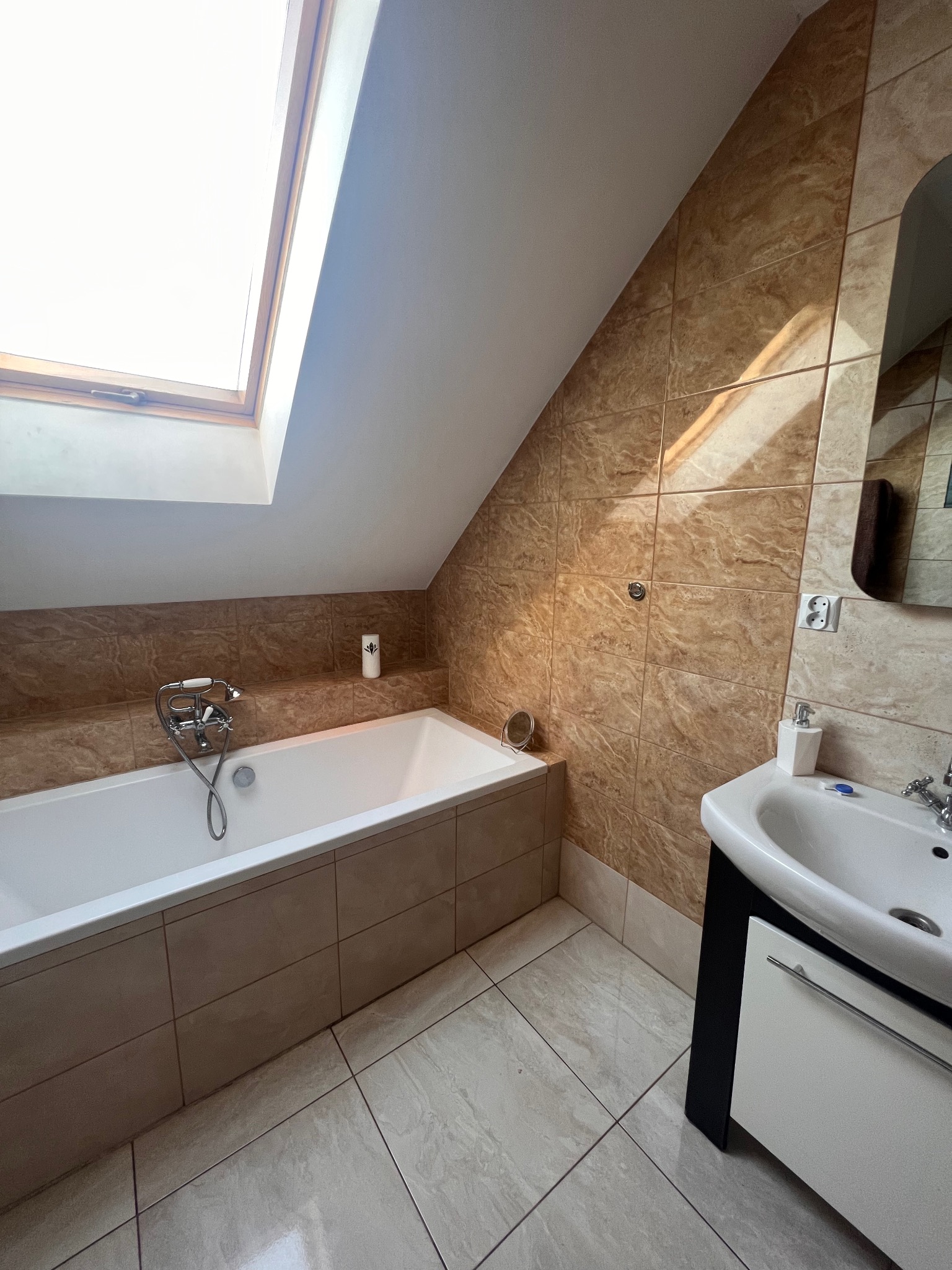 Ukończona łazienka na poddaszu z beżowymi płytkami imitującymi marmur, białą wanną pod oknem dachowym, umywalką z szafką i lustrem.
