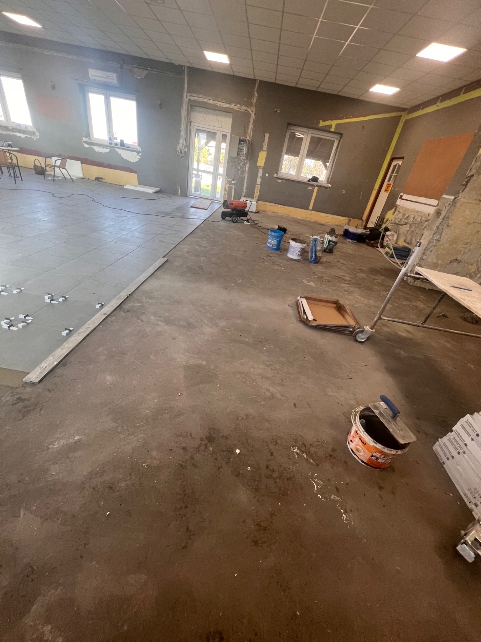 Wnętrze w trakcie układania płytek podłogowych, widoczne narzędzia glazurnicze, poziomica, krzyżyki dystansowe i wiadro z klejem. Część podłogi już wyłożona szarymi płytkami.