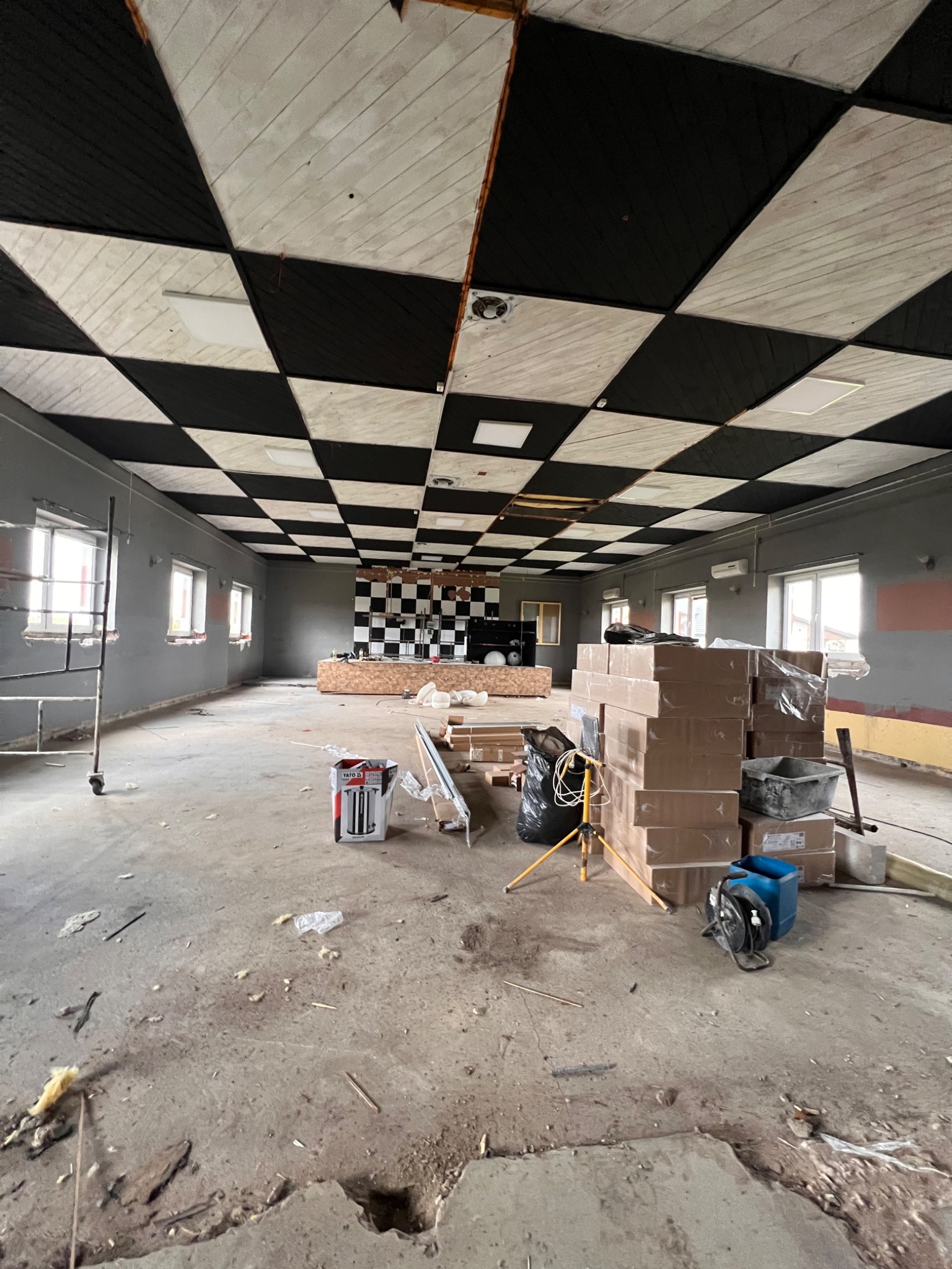 Przestronne wnętrze w trakcie prac wykończeniowych z szachownicą na suficie, widoczne materiały budowlane, drabina, narzędzia i surowy beton na podłodze.