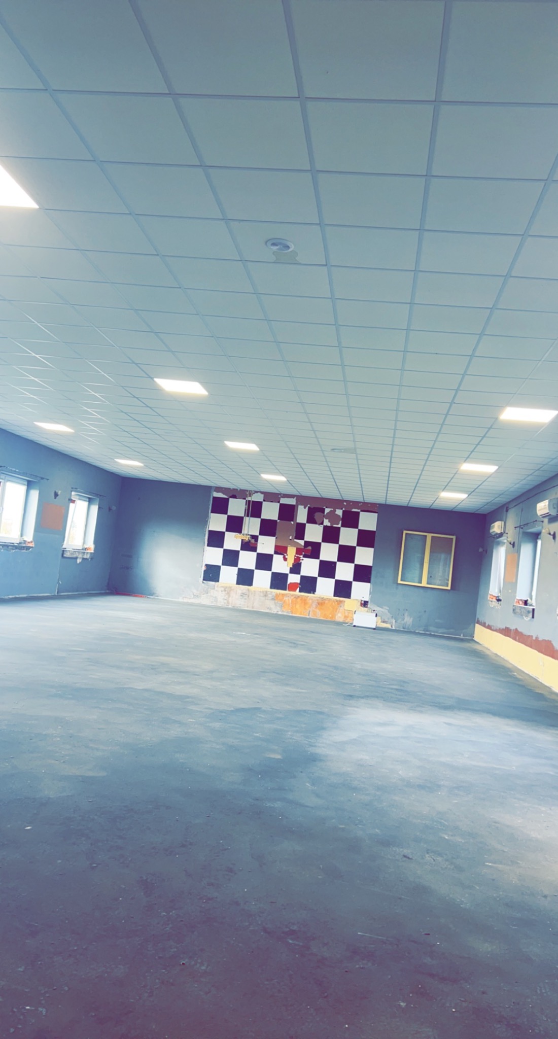 Wnętrze remontowanego pomieszczenia biurowego z szarą podłogą, ścianami w kolorze szarym i białą szachownicą na jednej ze ścian. Widoczne okna i sufit kasetonowy z oświetleniem.