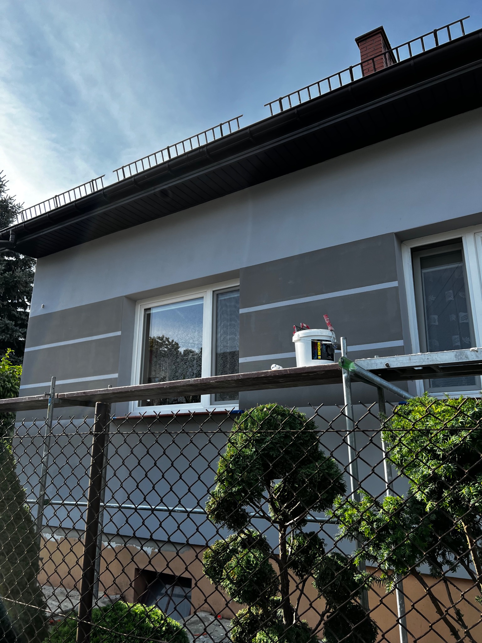 Szare wykończenie elewacji domu z poziomymi pasami, widoczne rusztowanie z wiadrem farby i pędzlami, ujęcie zza ogrodzenia z siatki.