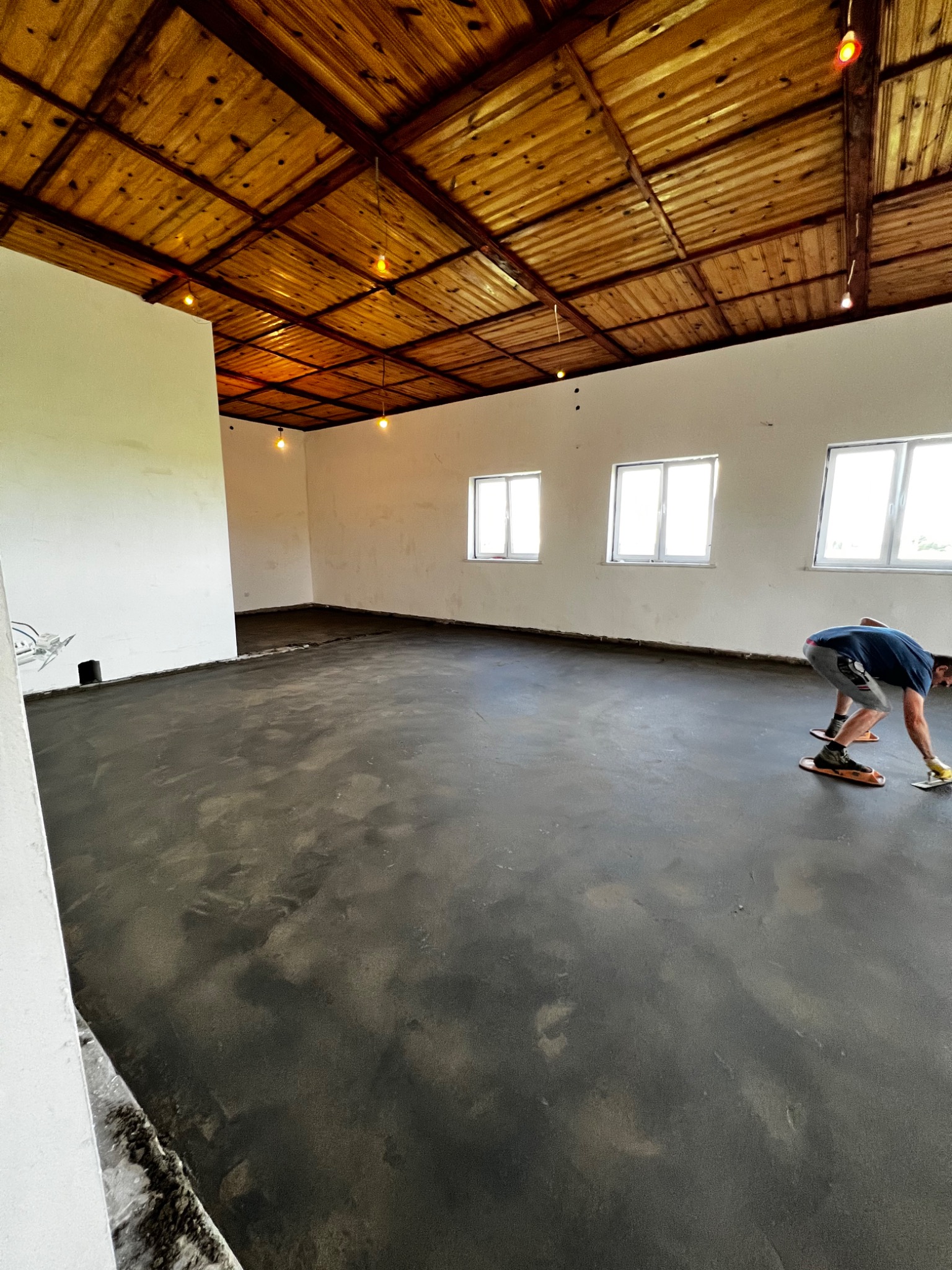 Wyrównywanie wylewki samopoziomującej w pomieszczeniu w Sierpcu, widoczne drewniane deski na suficie i pracownik w specjalnych nakładkach na buty.
