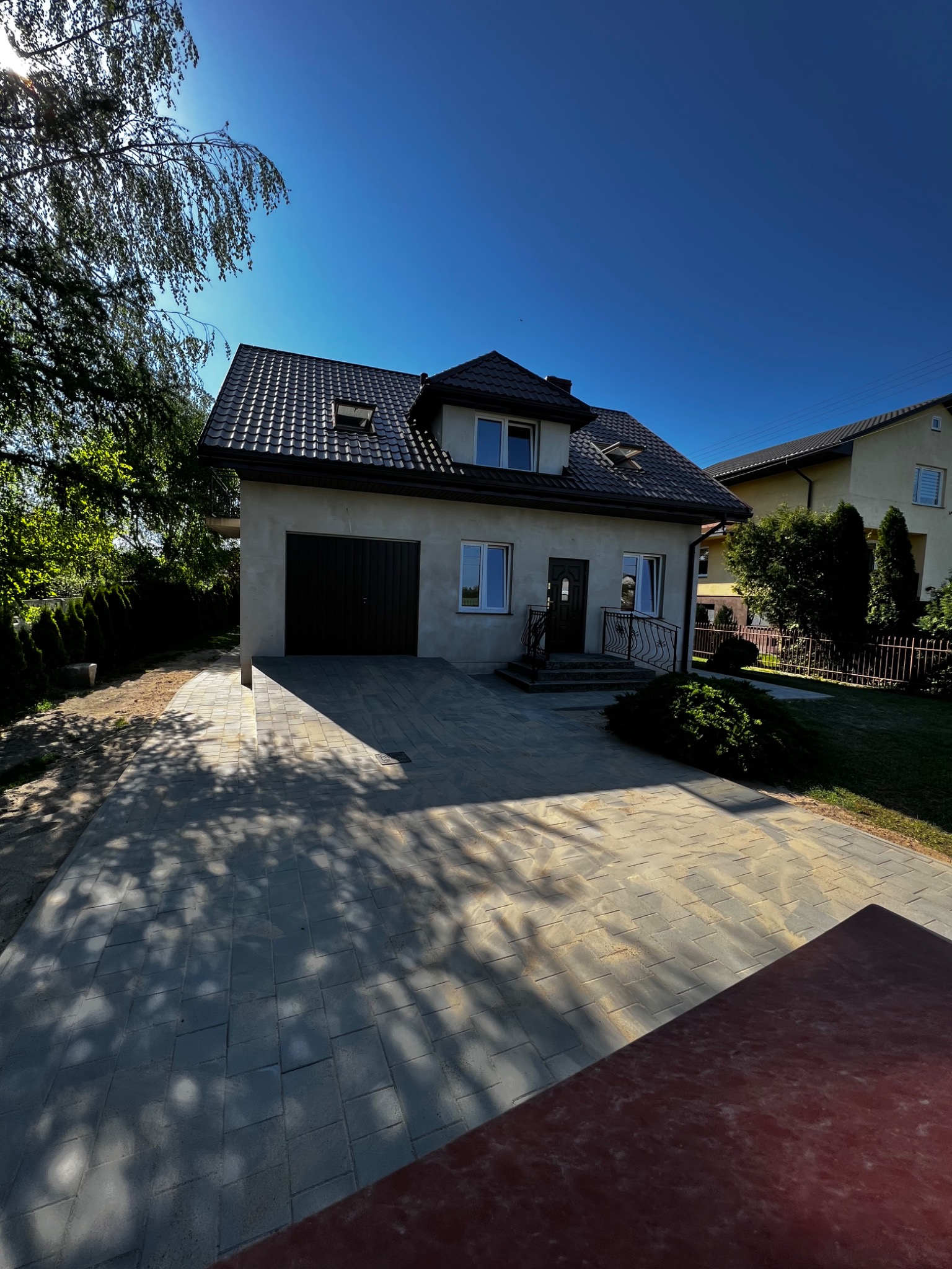 Dwukondygnacyjny dom jednorodzinny z garażem i podjazdem wyłożonym szarą kostką brukową, oświetlony mocnym słońcem, z widocznymi cieniami drzew.