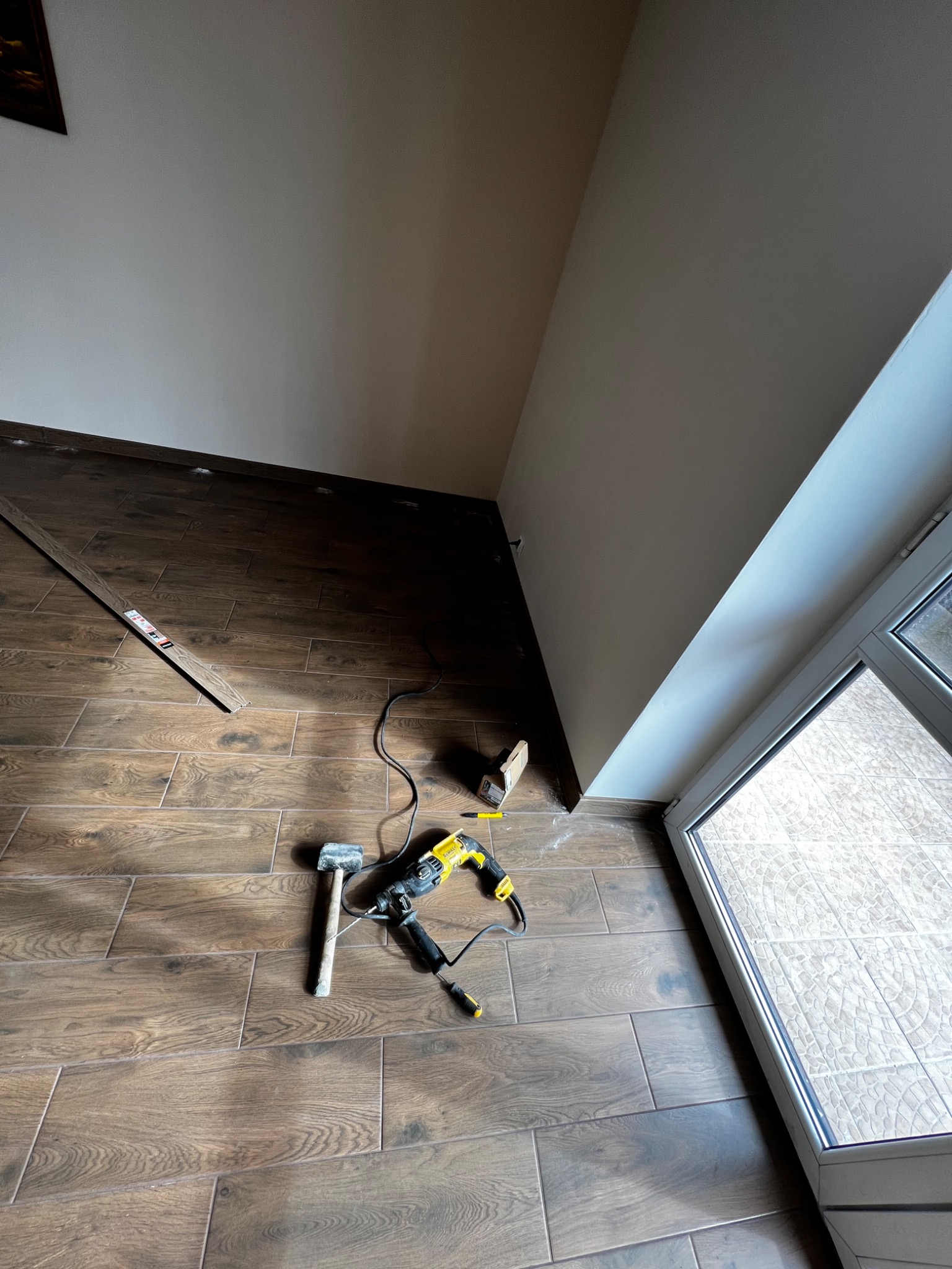 Narzędzia (wiertarka, młotek) na nowo położonej podłodze z płytek imitujących drewno, widoczny fragment listwy przypodłogowej i okno balkonowe.