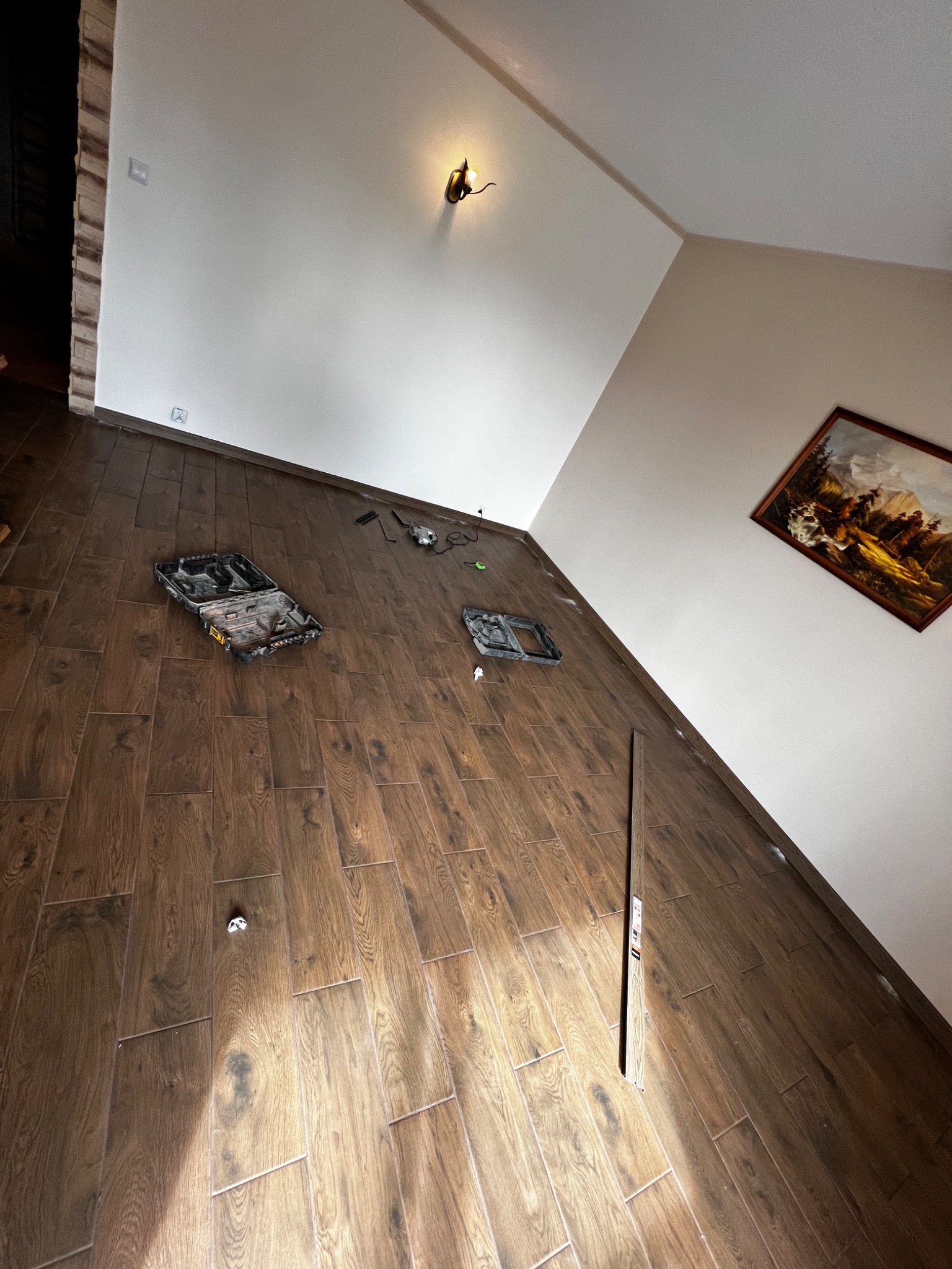 Ukośnie ułożone, brązowe płytki podłogowe imitujące drewno, z rozłożonymi narzędziami i otwartą skrzynką narzędziową, widoczne fragmenty białych ścian i obrazu wiszącego na ścianie.