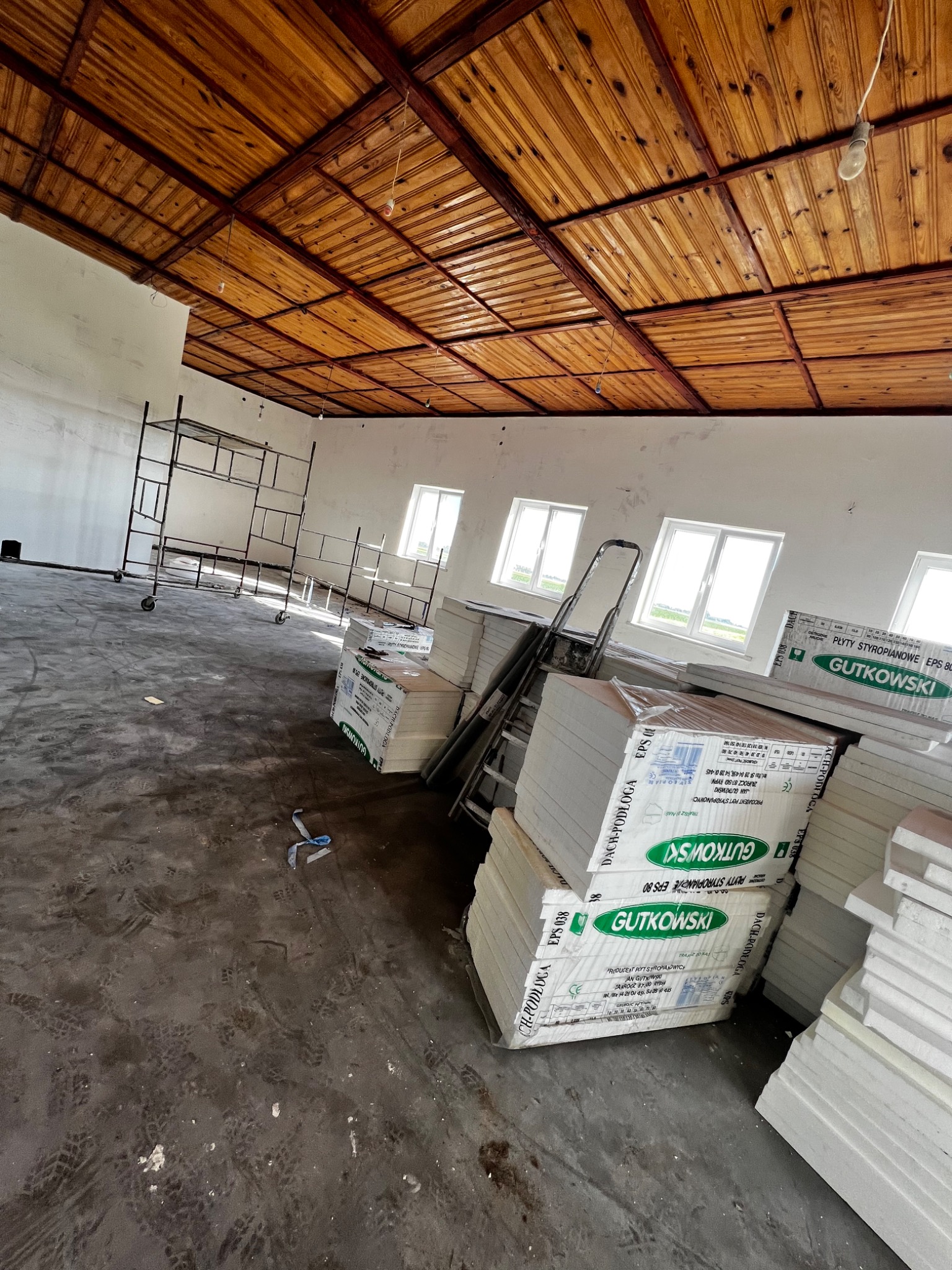Styropianowe płyty izolacyjne Gutkowski w opakowaniach, ustawione w stosy w surowym wnętrzu z drewnianym sufitem i rusztowaniem, przygotowane do ocieplenia.