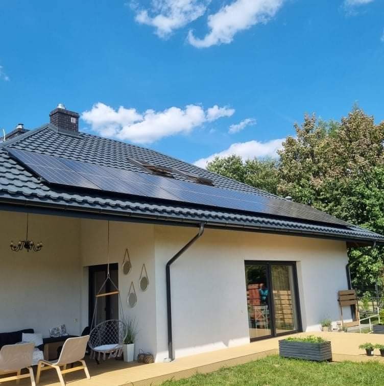 Dom jednorodzinny z panelami słonecznymi na dachu, widok z ogrodu w słoneczny dzień, dekoracyjne elementy na elewacji, ciemna dachówka.