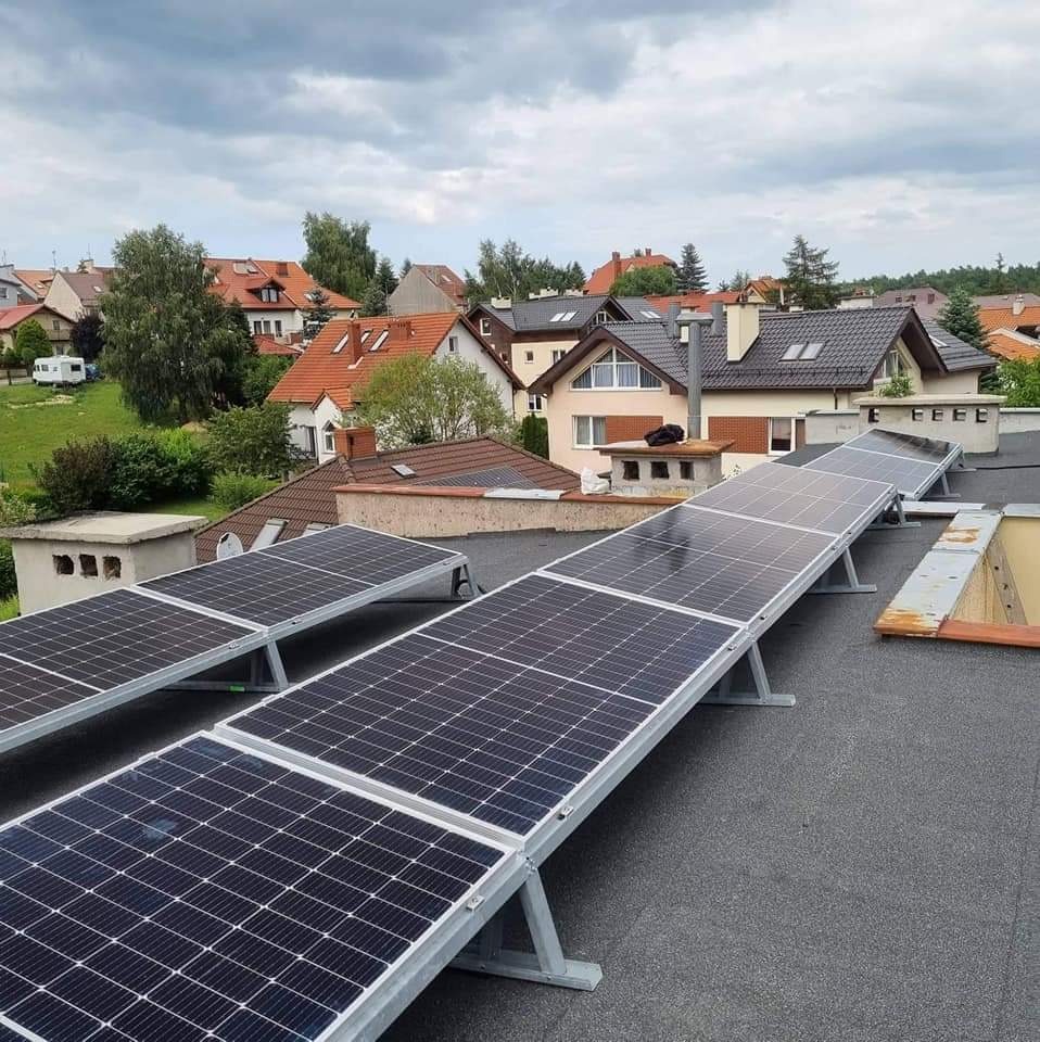 Rząd paneli słonecznych zamontowanych na płaskim dachu pokrytym papą, z widokiem na okoliczne domy i pochmurne niebo.