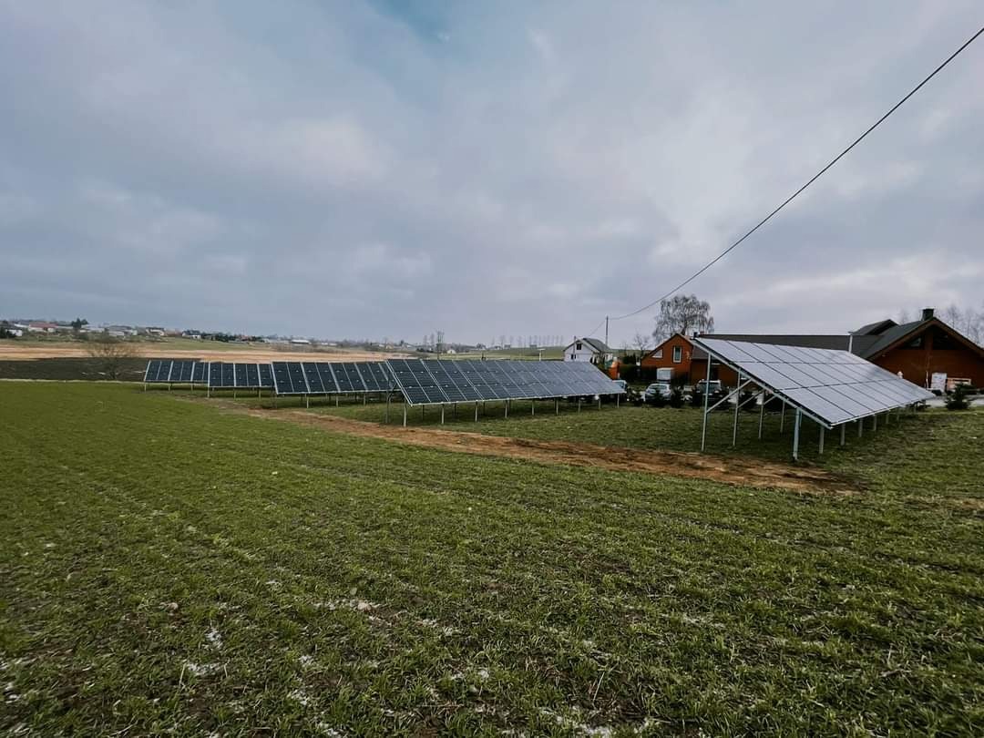 Rozległa instalacja fotowoltaiczna na gruncie rolnym, panele ustawione rzędami, w tle domy jednorodzinne pod pochmurnym niebem.