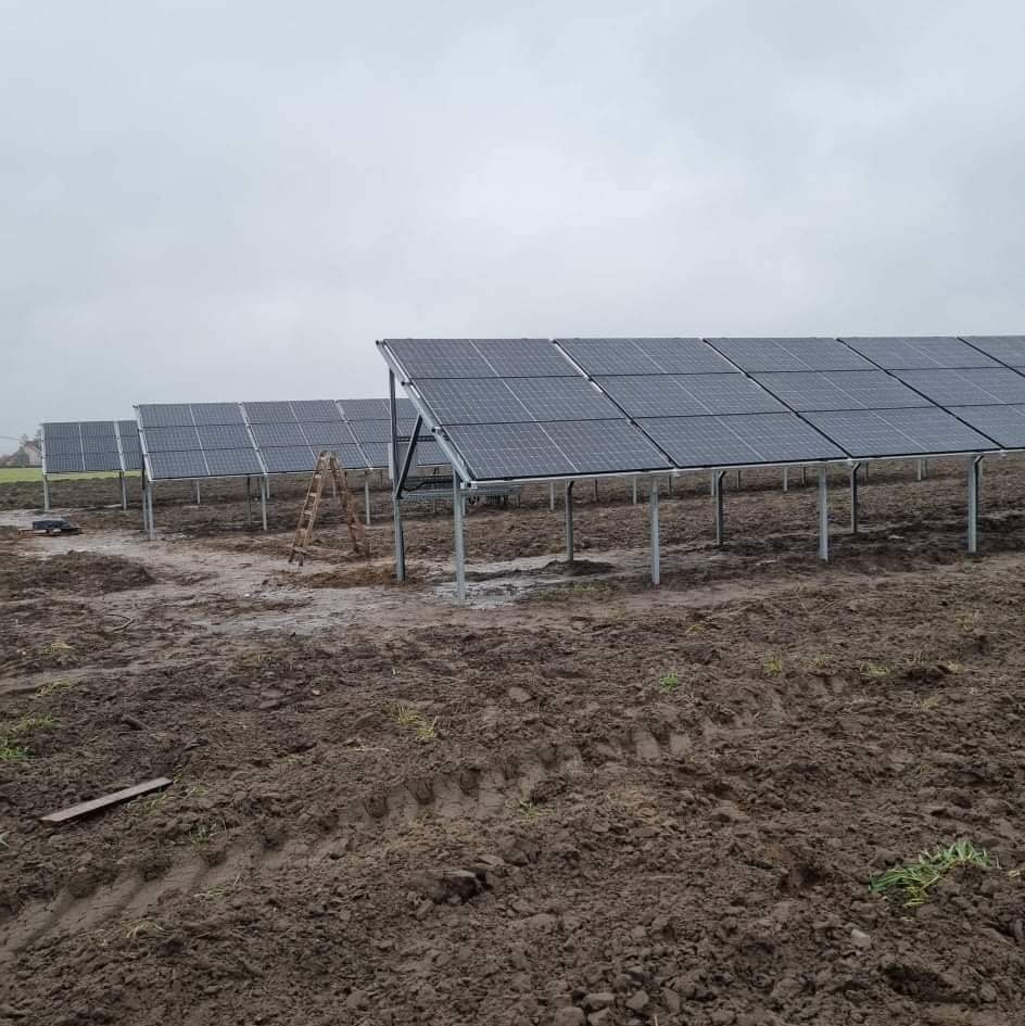 Montaż paneli słonecznych na polanie z błotnistym podłożem, widoczna drabina oparta o konstrukcję, pochmurne niebo.