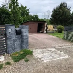 Nowoczesna brama posesji z szarych, betonowych bloczków z czarną skrzynką na listy i domofonem, prowadząca na podjazd z kostki brukowej i widocznym garażem blaszanym w tle.