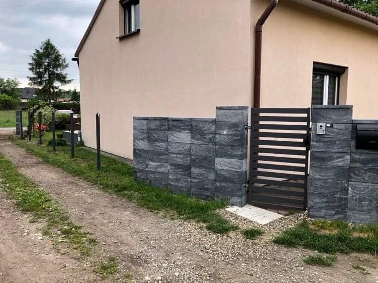 Nowoczesna brama posesyjna z szarych, betonowych bloczków imitujących kamień, z czarnymi, poziomymi przęsłami, zamontowana przy domu z beżową elewacją. Widoczny domofon i skrzynka na listy. Ścieżka...