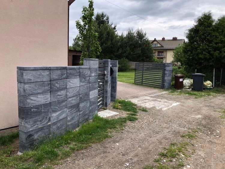 Nowoczesne ogrodzenie posesji z szarych, betonowych bloczków z poziomymi przęsłami, widok na podjazd i dom w tle.