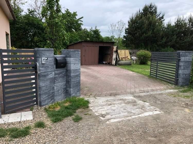 Nowoczesna brama posesji z szarych, betonowych bloczków z czarną skrzynką na listy i domofonem, prowadząca na podjazd z kostki brukowej i widocznym garażem blaszanym w tle.