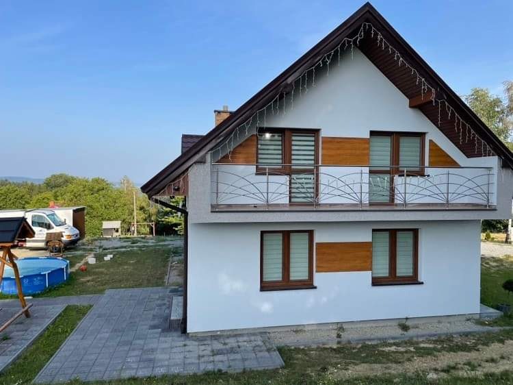 Dwukondygnacyjny dom z białymi ścianami, brązowym dachem i dekoracyjnymi lampkami, z balkonem z metalową balustradą i drewnianymi akcentami wokół okien, na tle zielonego ogrodu i błękitnego nieba.